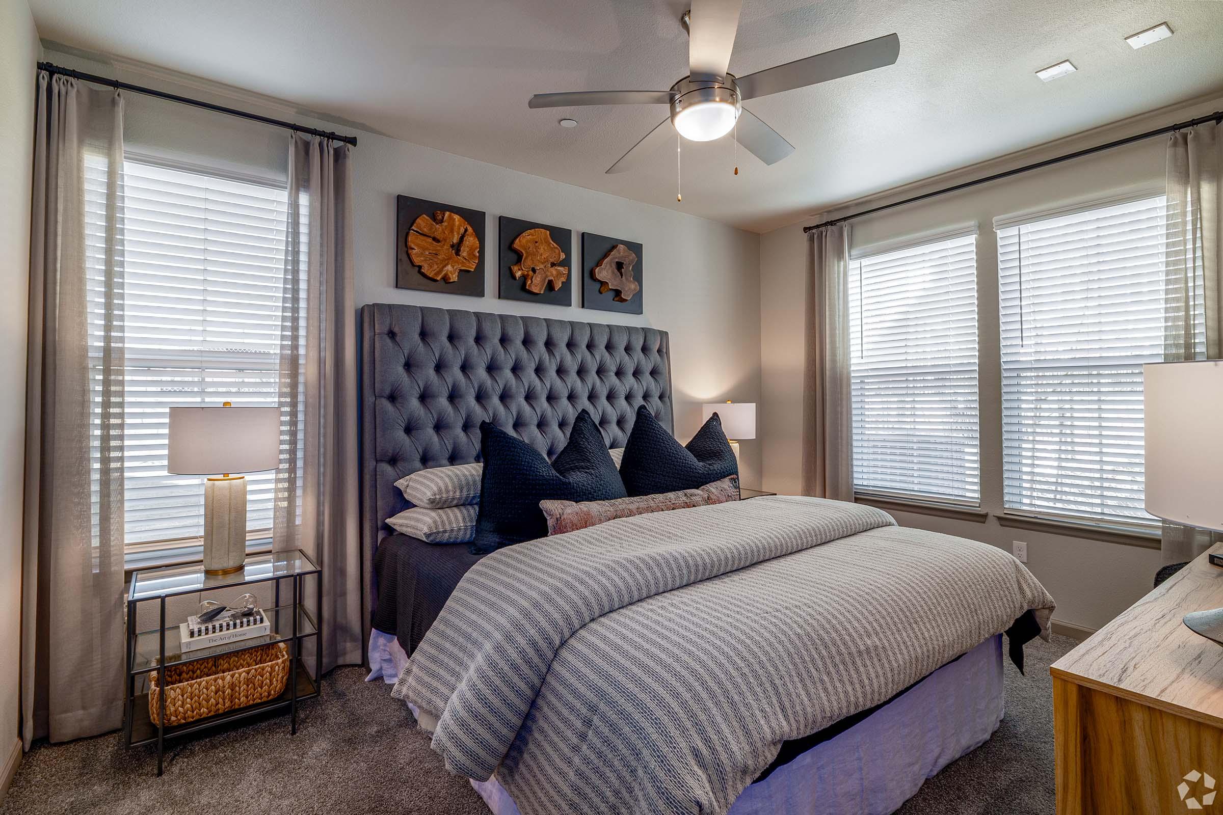 A stylish bedroom featuring a tufted gray upholstered bed with decorative pillows, a nightstand with a lamp, and large windows dressed with sheer curtains. Art pieces hang above the headboard, and a ceiling fan adds a modern touch to the cozy atmosphere. A wooden dresser completes the room's elegant design.