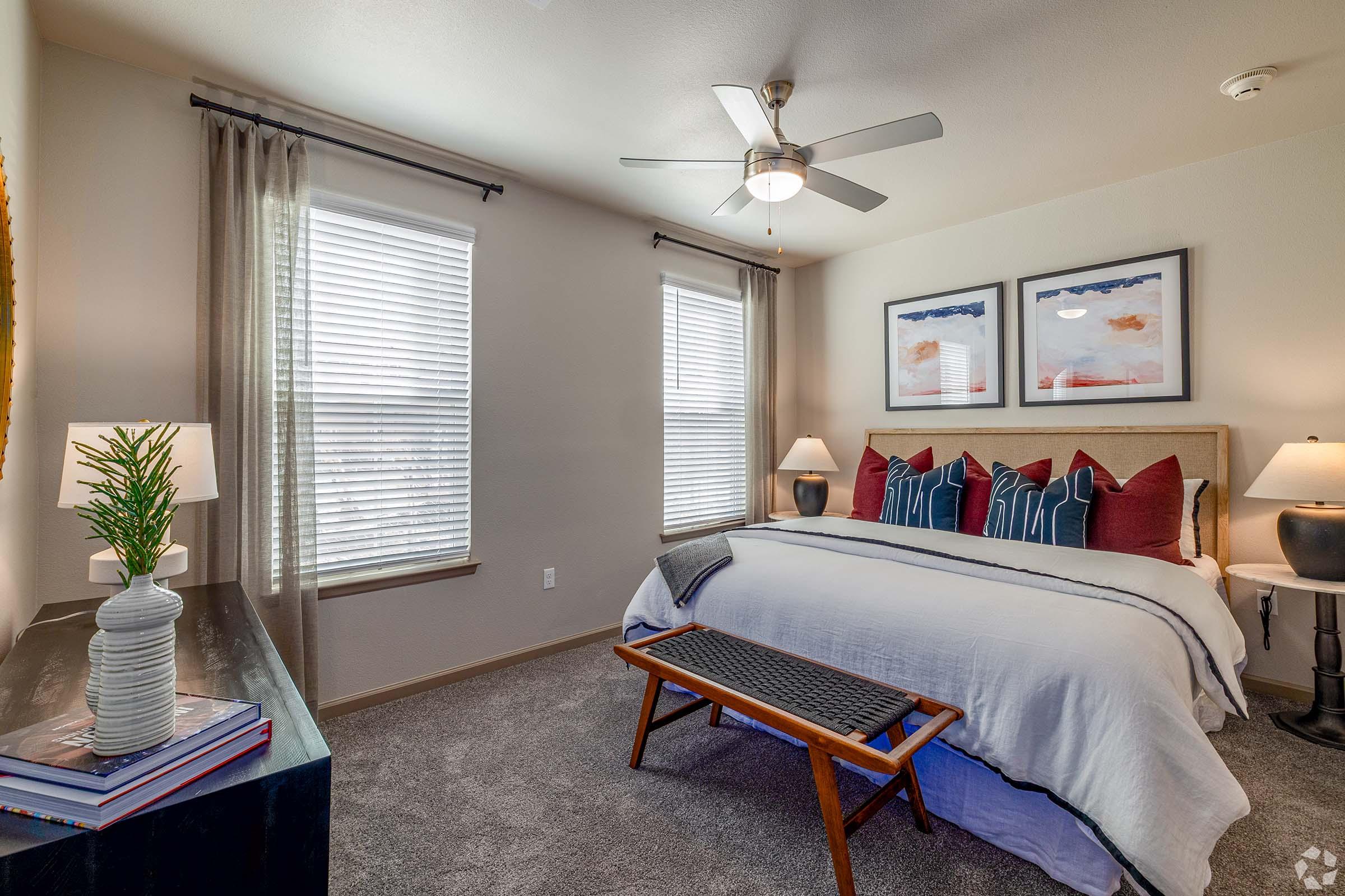 A cozy bedroom featuring a queen-sized bed with decorative throw pillows, a wooden bench at the foot of the bed, a stylish nightstand with lamps, and two windows with blinds allowing natural light. The walls display framed artwork, and a small plant adds a touch of greenery. The room has a warm and inviting atmosphere.