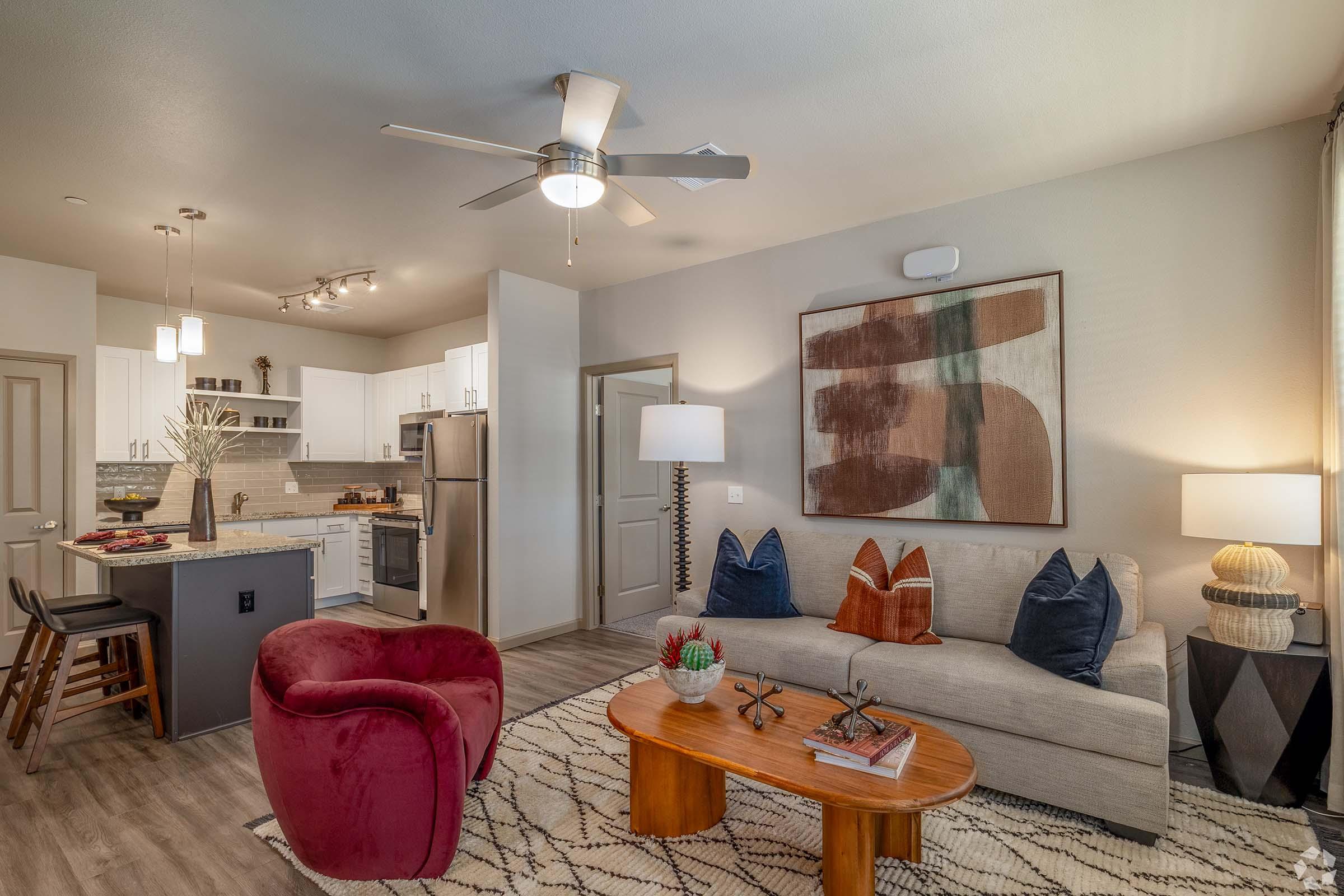 A modern living room featuring a light gray sofa with colorful throw pillows, a red accent chair, a wooden coffee table, and a patterned rug. In the background, there is a dining area with a kitchen visible, showcasing white cabinets and stainless steel appliances. The space is well-lit with a ceiling fan and stylish decor.