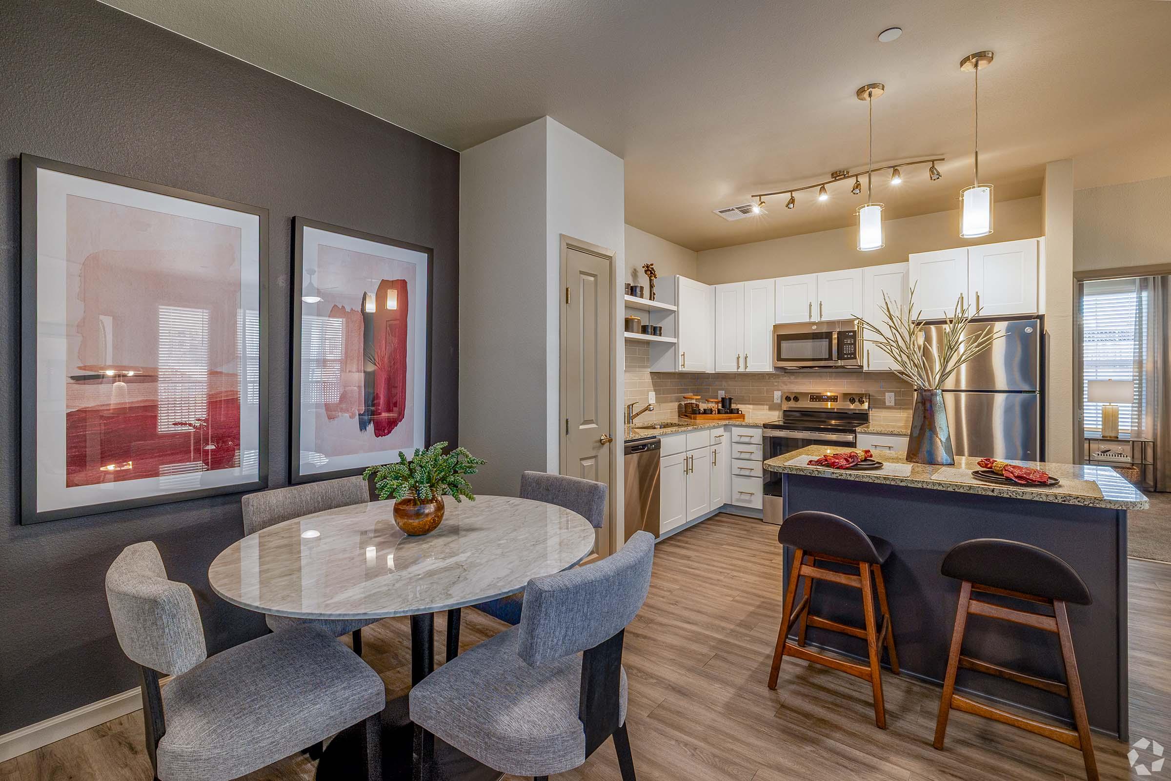 A modern kitchen and dining area featuring a round marble table with gray chairs, a contemporary kitchen with white cabinets, stainless steel appliances, and wooden bar stools. The walls are decorated with abstract art, and the space is well-lit with natural light streaming in.