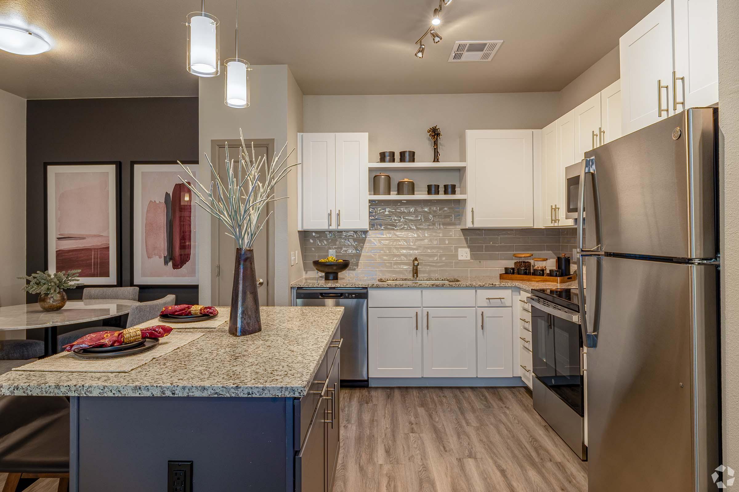Modern kitchen featuring light-colored cabinets, stainless steel appliances, and a granite countertop. The space includes decorative elements like a vase with branches, stylish dishes, and wall art. A dining table is visible in the background, creating a welcoming and contemporary atmosphere.