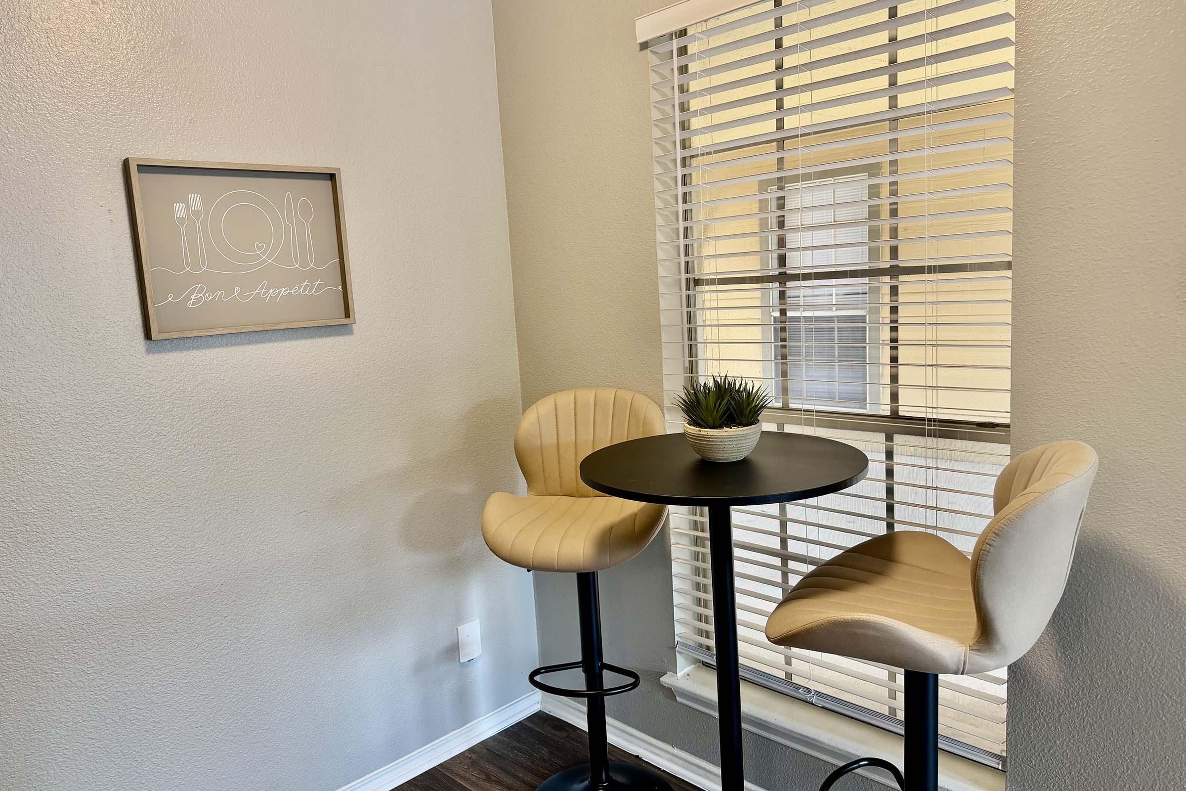 A cozy dining nook featuring a small black round table with two light beige bar stools. A potted plant sits on the table, and the wall has a framed decorative piece with a minimalist design of utensils. Natural light streams in through a window with blinds.