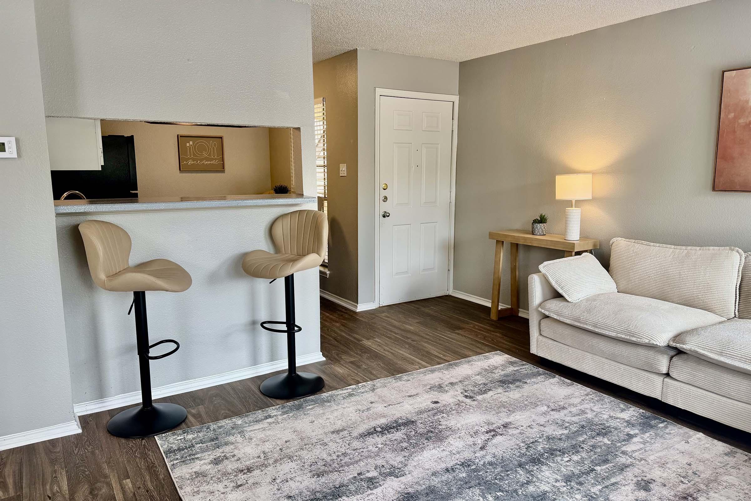 A cozy living space featuring a light-colored sofa, a small wooden table, and two modern bar stools. The walls are painted in a soft gray, and there’s a door leading outside along with a lamp providing warm light. A rug adds texture to the floor, creating a welcoming atmosphere.