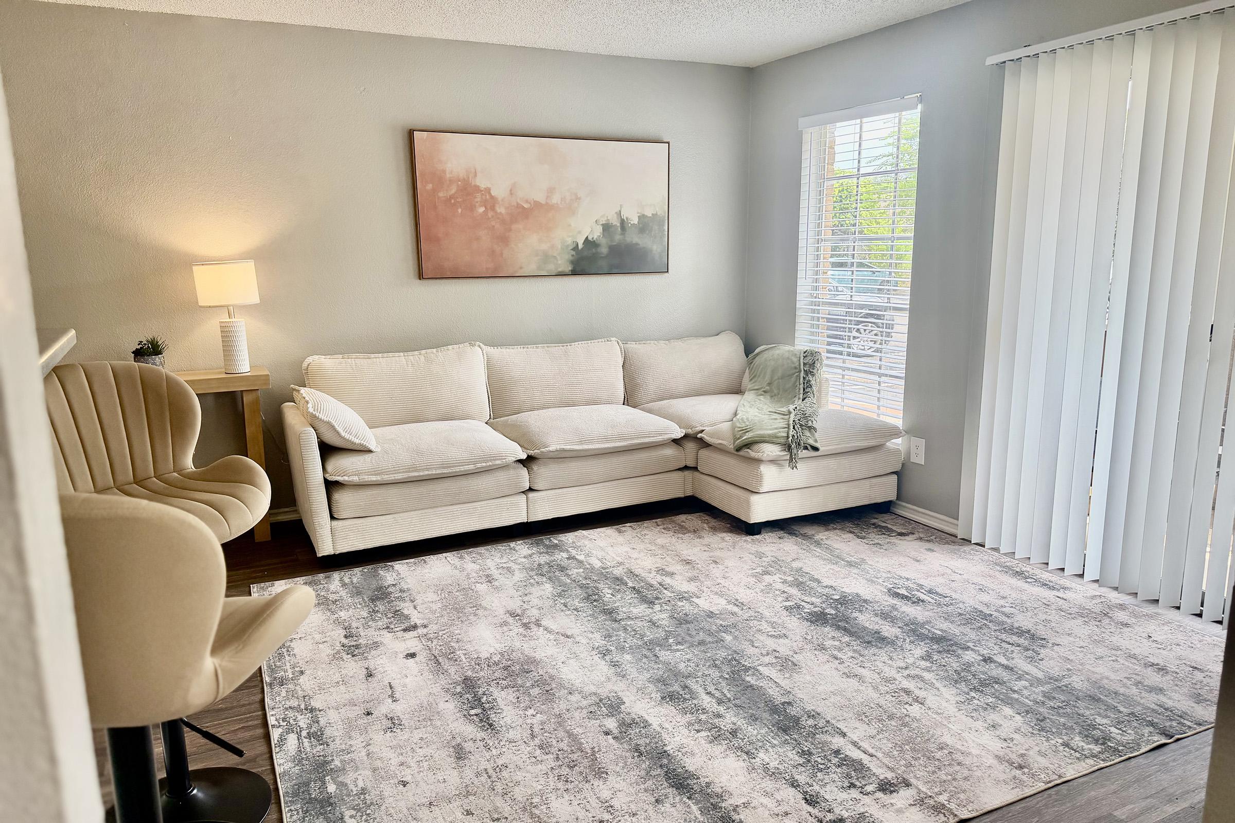 A cozy living room featuring a light beige sectional couch with a green throw blanket, a soft area rug, a modern lamp on a wooden side table, and an accent chair. Natural light filters through white vertical blinds, enhancing the tranquil ambiance of the space. A serene abstract painting hangs on the wall.