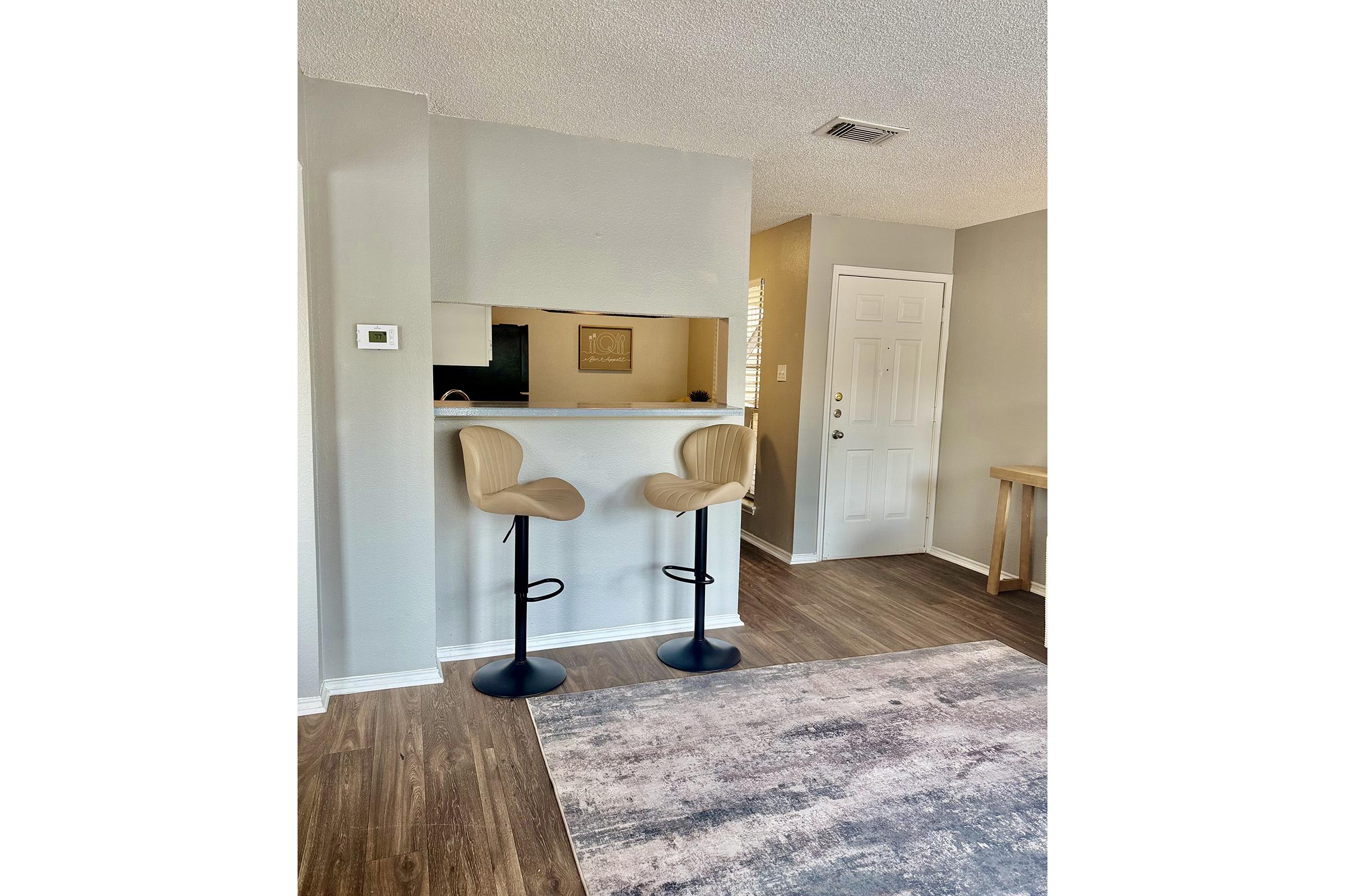 A modern interior view of a room featuring two minimalist bar stools at a counter. The floor is wooden, and a soft rug lies underneath. A door is visible to the right, and the walls are painted in a light gray color. The area appears bright and inviting, with a casual, contemporary vibe.