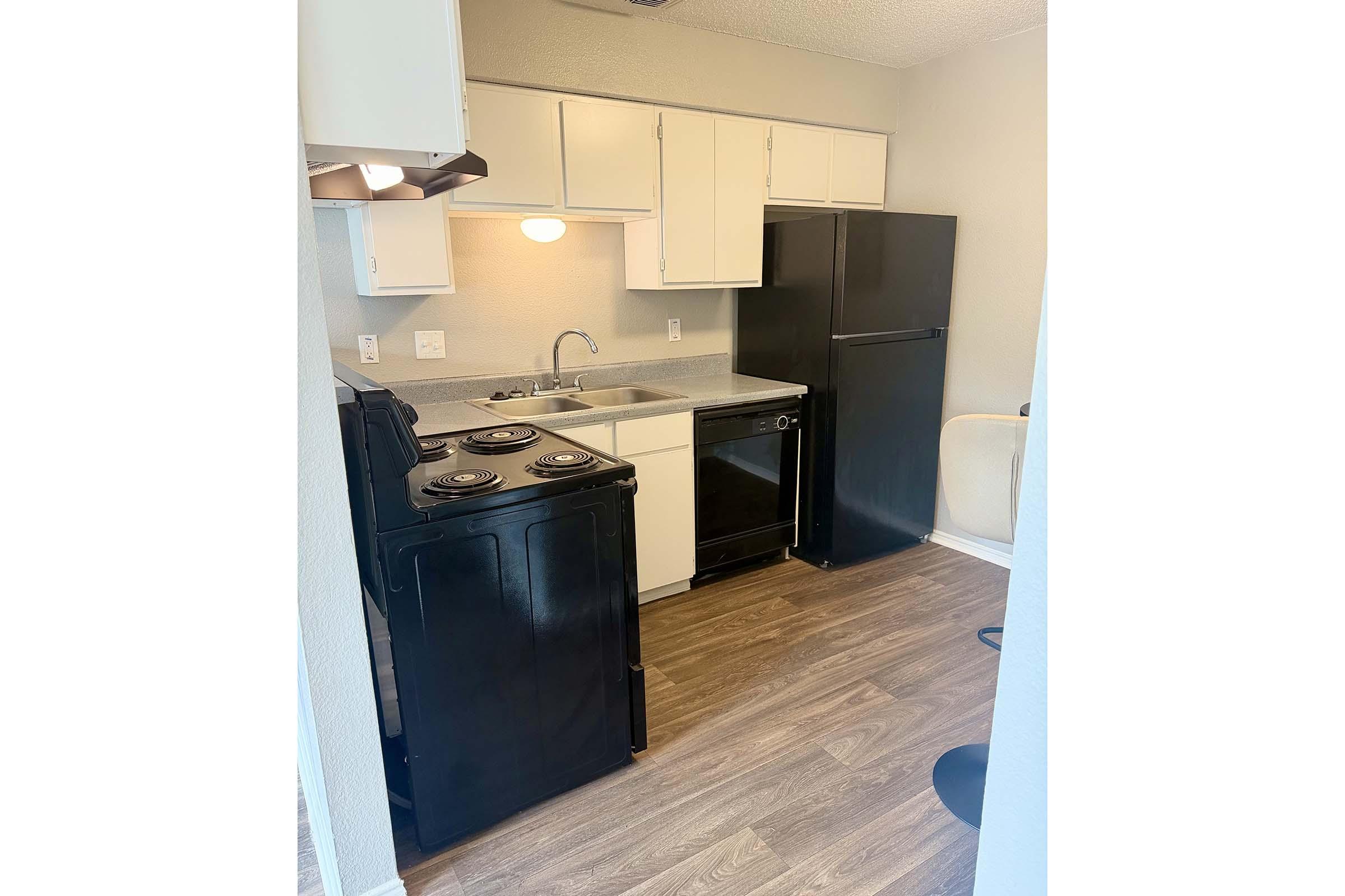 A small kitchen featuring black appliances, including a stove, oven, and refrigerator. The countertop is light-colored with a sink and cupboard space above. The floor has a wood-like finish, and there is a small dining area visible with two stools. The overall design is simple and functional.
