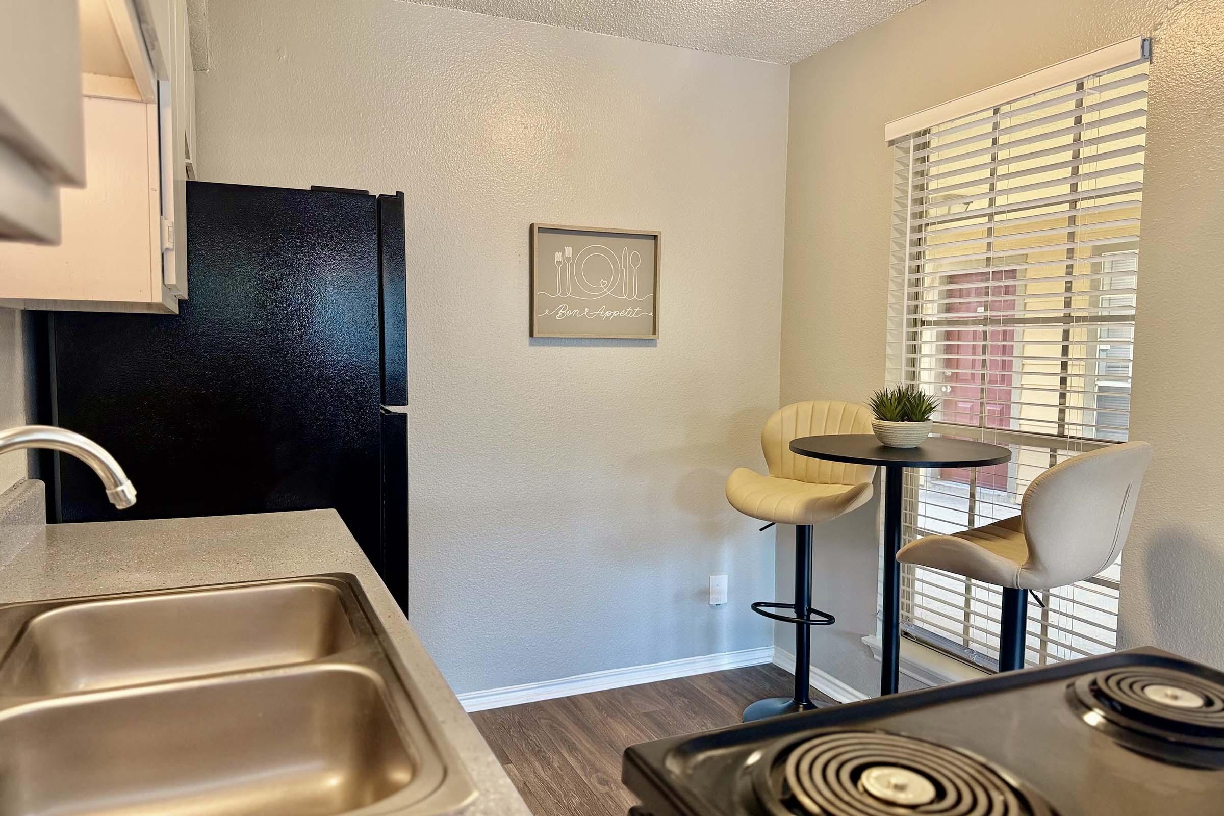 A compact kitchen featuring a black refrigerator and stove, a gray countertop, and a small dining area with two bar stools and a round table. The wall has a framed decoration, and there's a potted plant on the table. Natural light filters through the blinds, creating a cozy ambiance.