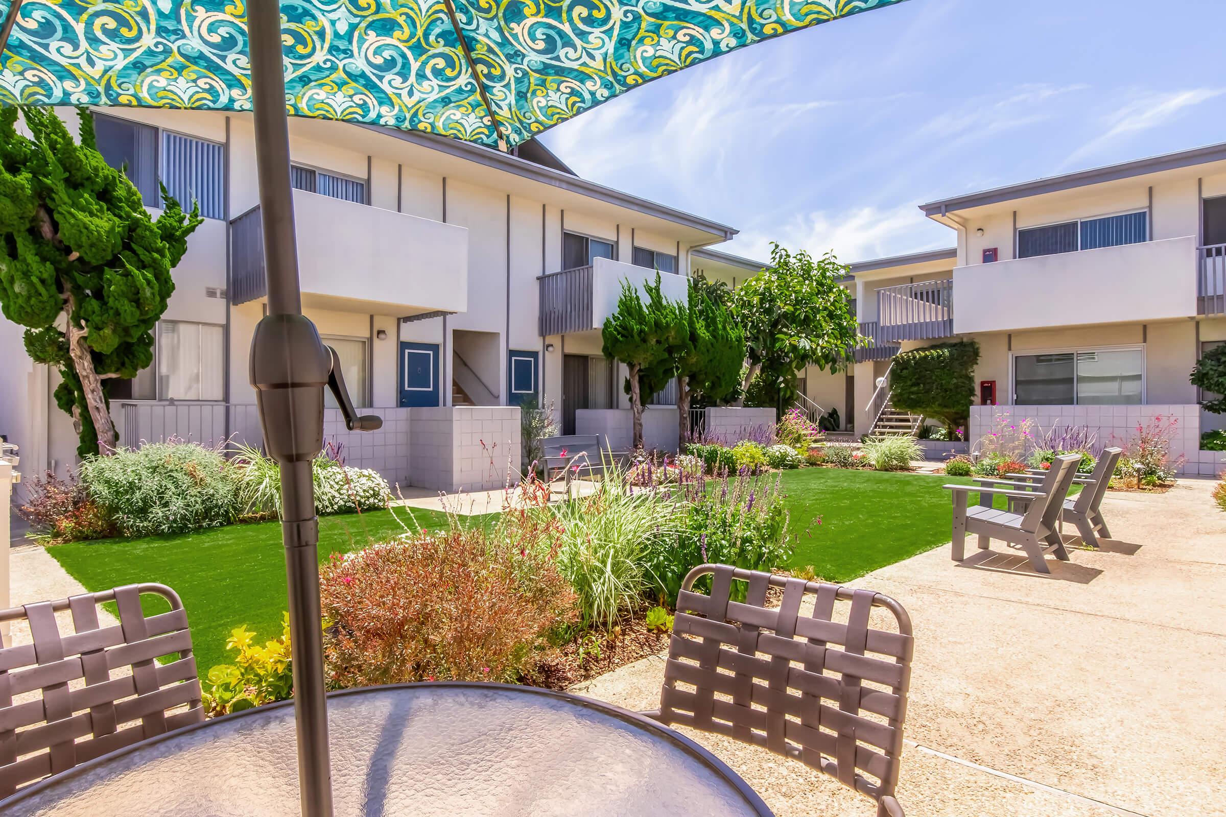 A sunny courtyard featuring landscaped gardens with colorful plants, surrounded by modern buildings. There are comfortable seating areas with patio chairs and a table under a patterned umbrella, providing a relaxing outdoor space. The atmosphere is bright and inviting, typical of a well-maintained residential complex.