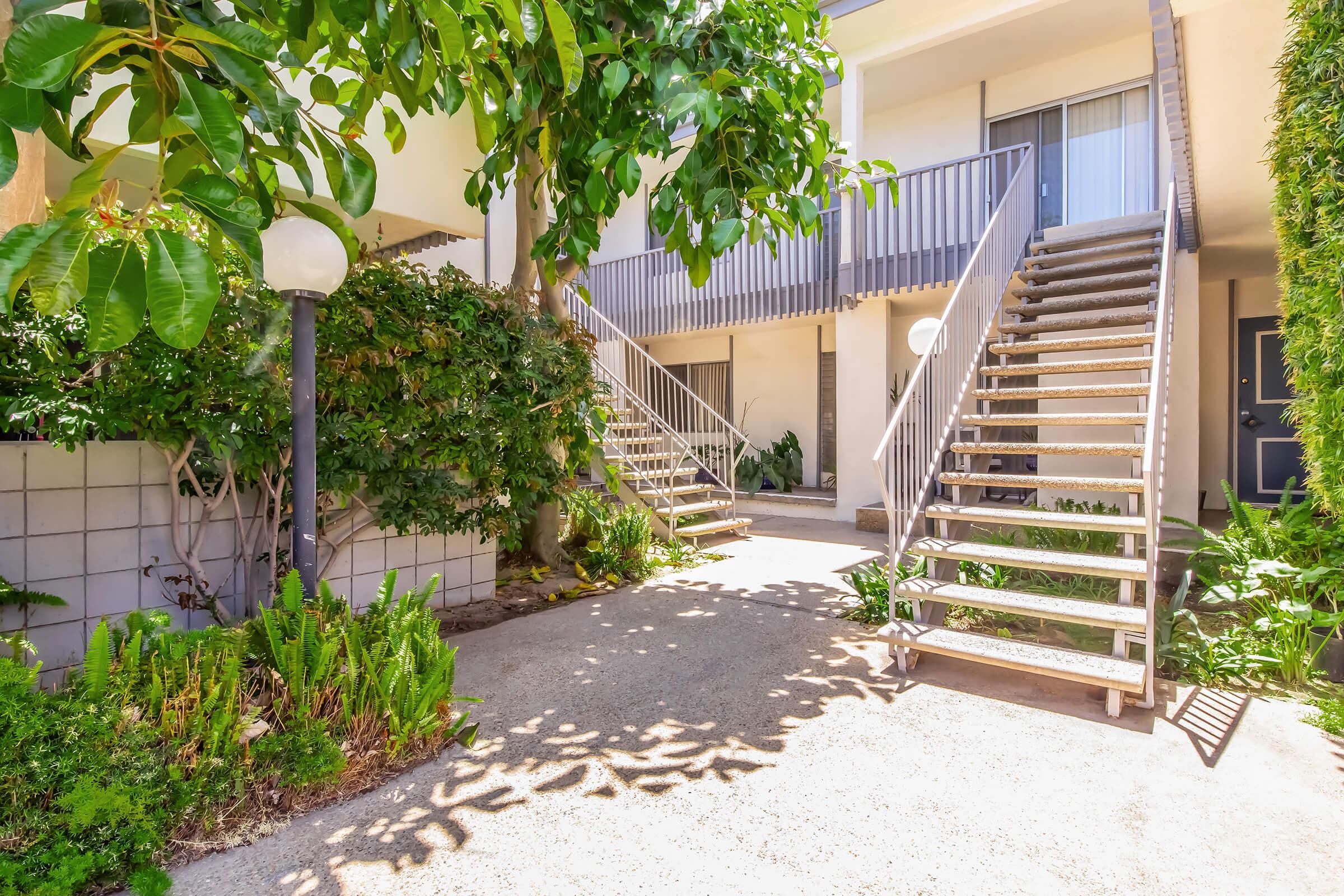 A well-lit outdoor area featuring two sets of metal stairs leading to upper units, surrounded by lush greenery and small plants. The pathway is paved, and there are lampposts offering illumination. The setting appears inviting and well-maintained, typical of a residential complex.