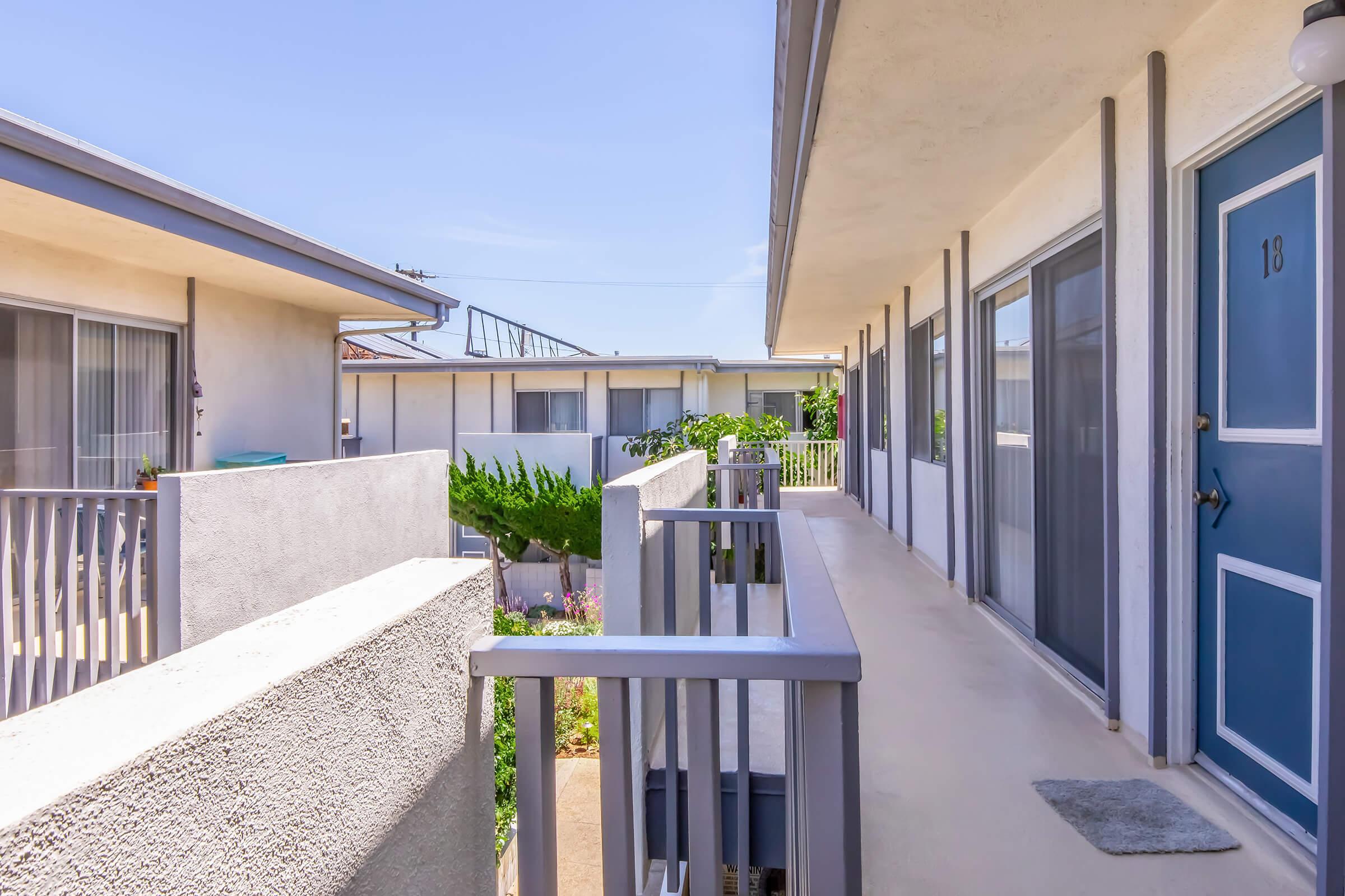 View of a balcony corridor in a multi-unit apartment complex. The scene features light-colored walls, blue doors, and plant greenery. The sky is clear, and the architecture includes railings and access points to individual units. The area appears well-maintained and inviting.