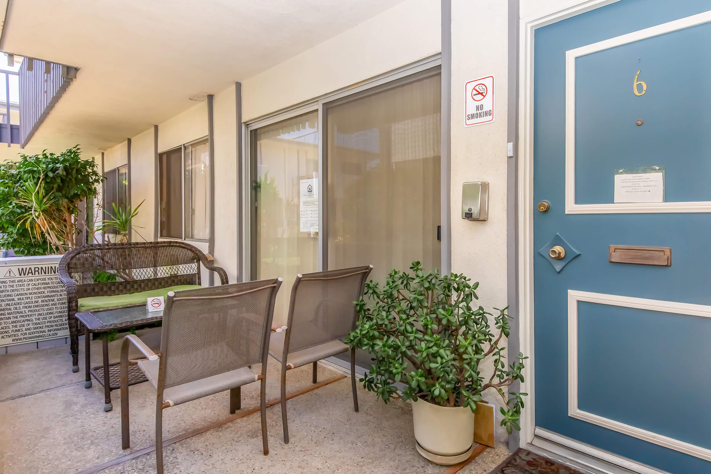 A cozy entryway with a blue door labeled "6," featuring a small seating area with two chairs and a table. Potted plants add greenery. A sign indicates no smoking in the area, and large windows allow natural light to illuminate the space.