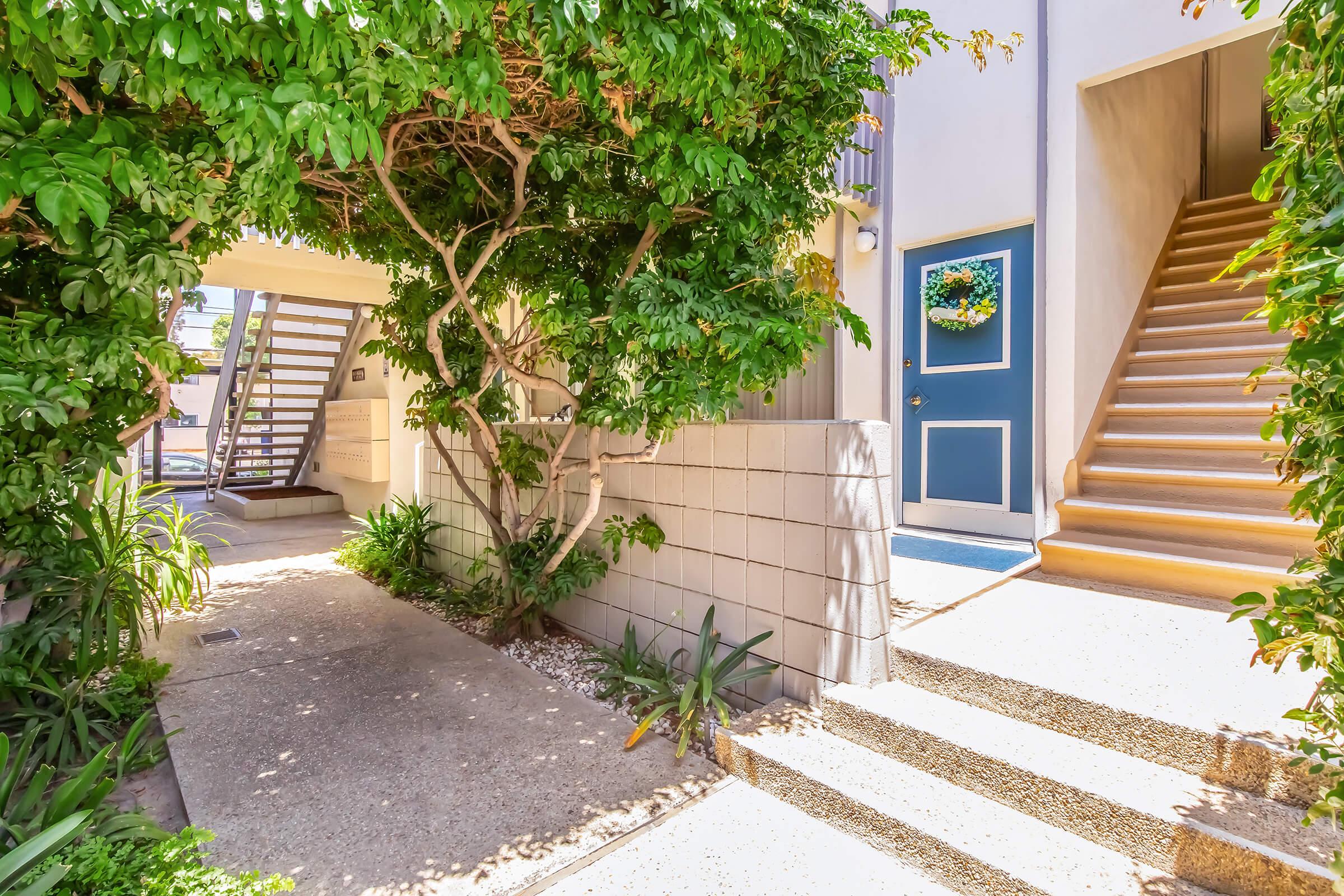 A well-lit entrance to a building, featuring a pathway flanked by lush greenery on both sides. On the left, a staircase leads to the upper floor, while the right side showcases a blue door adorned with a round wreath, giving a welcoming feel to the space.