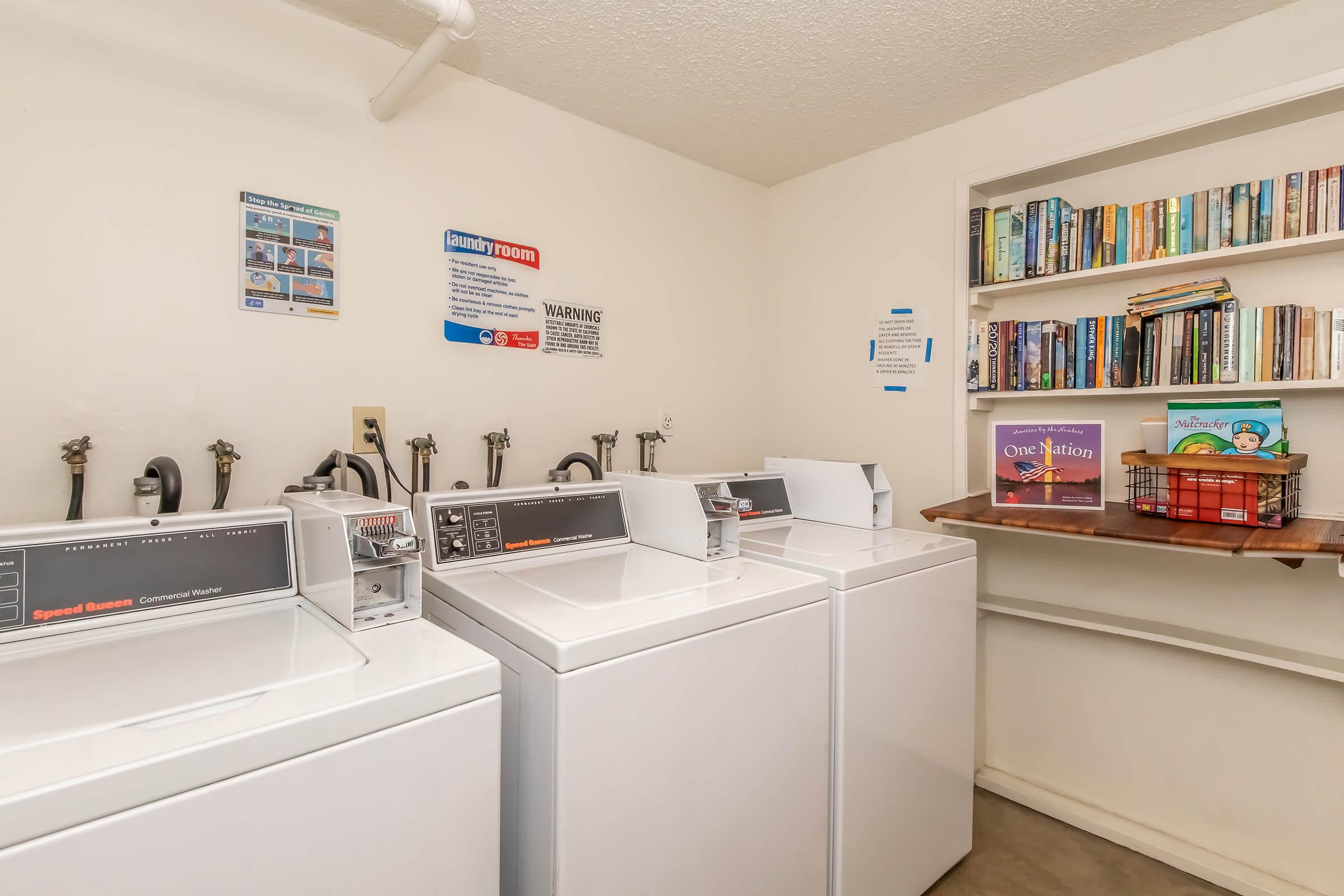 Laundry room featuring three white washing machines, a shelf filled with books on the wall, and a small basket. Informational signs about laundry usage are displayed nearby, creating a functional and organized space for laundry tasks.