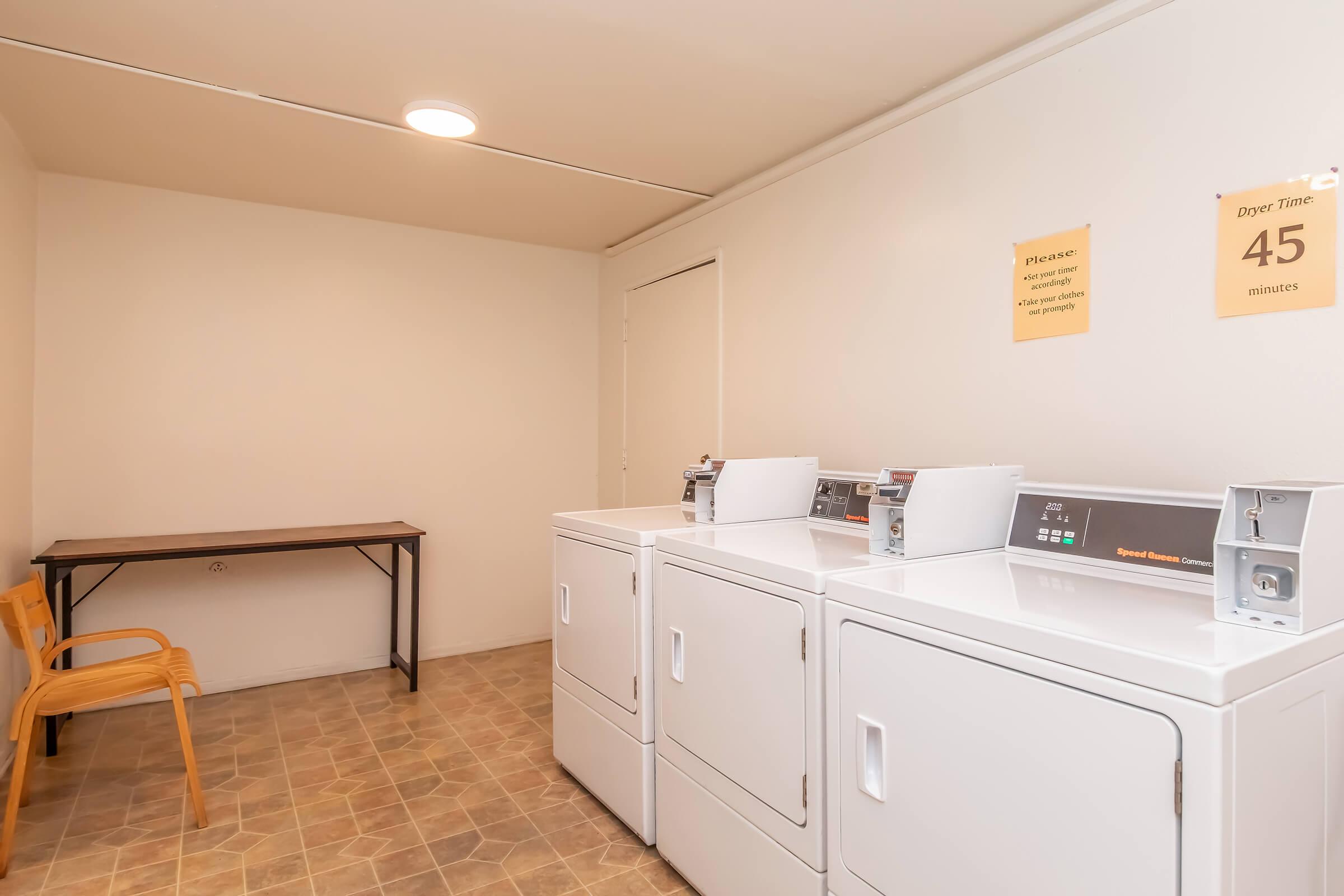 A clean and well-lit laundry room featuring three white washing machines arranged in a line. There is a small table and a chair in the room. On the wall, there are two yellow signs: one instructing users on laundry procedures and another indicating dryer time of 45 minutes. The floor is tiled.