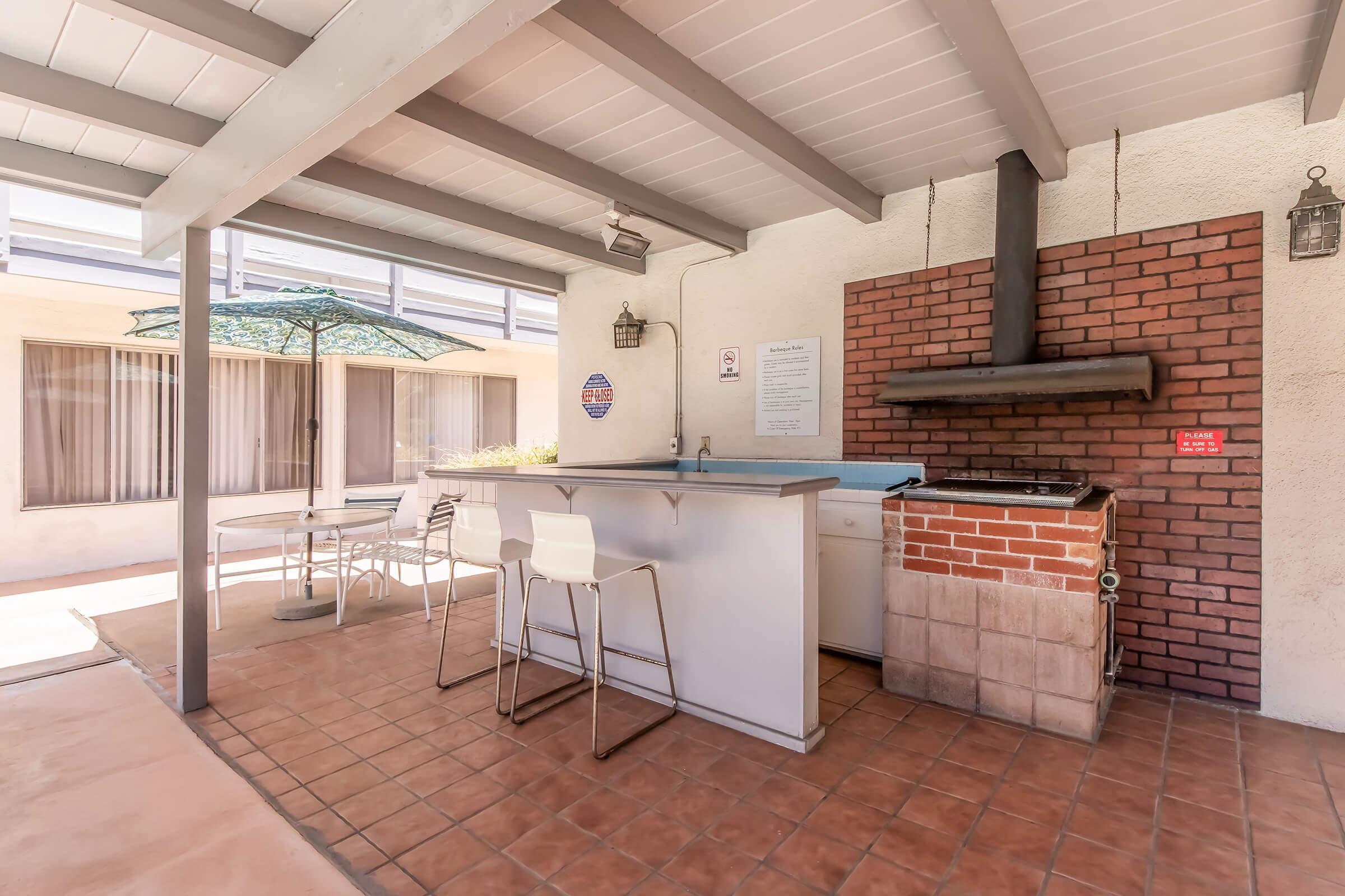 An outdoor patio area featuring a brick barbecue grill, a counter with bar stools, a small table and chairs, and an umbrella for shade. The space has tiled flooring and is surrounded by large windows.