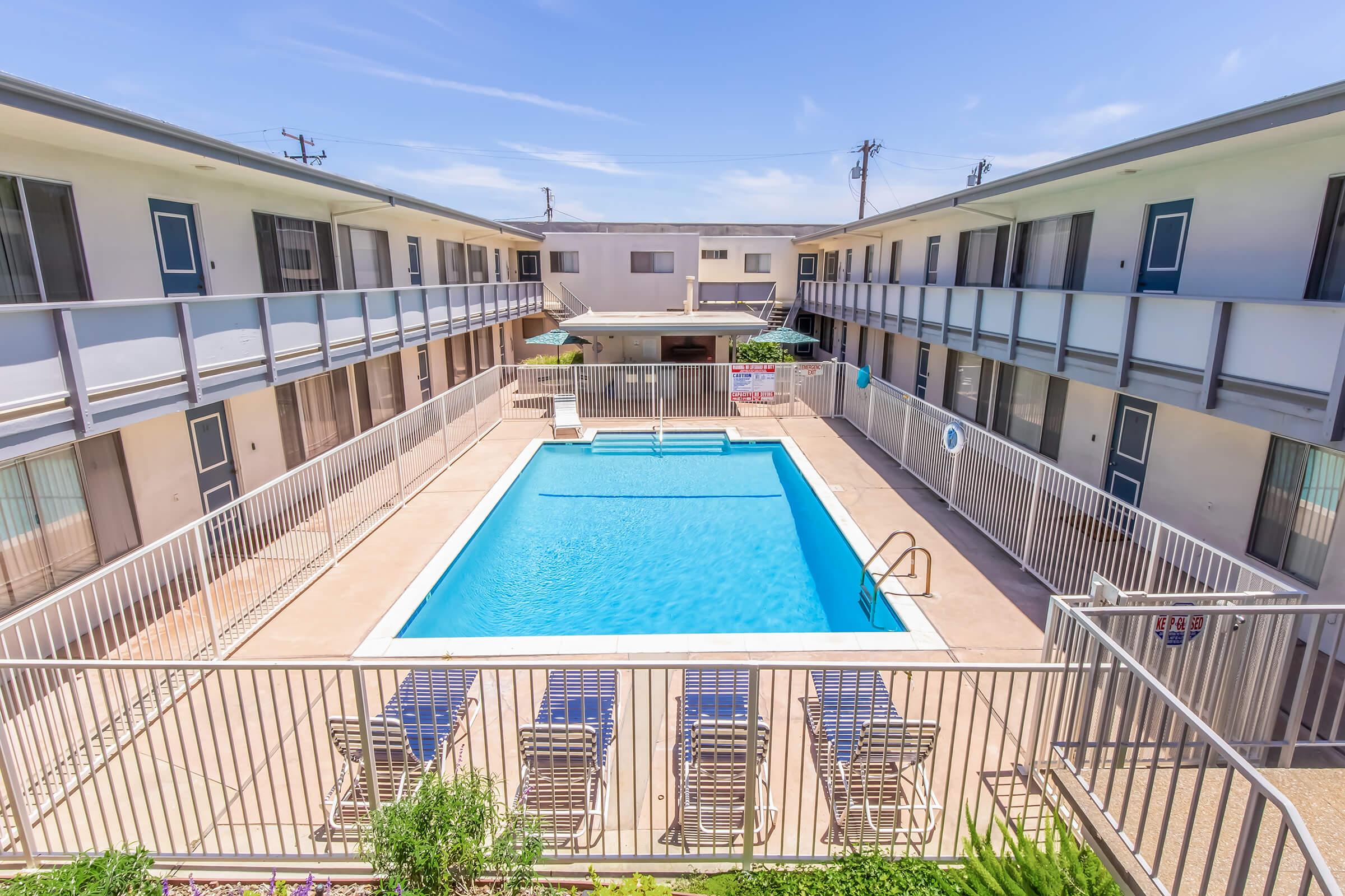 View of a rectangular swimming pool surrounded by lounge chairs, located in the center of an apartment complex. The two-story buildings have balconies overlooking the pool area, with clear blue skies above and surrounding fencing.
