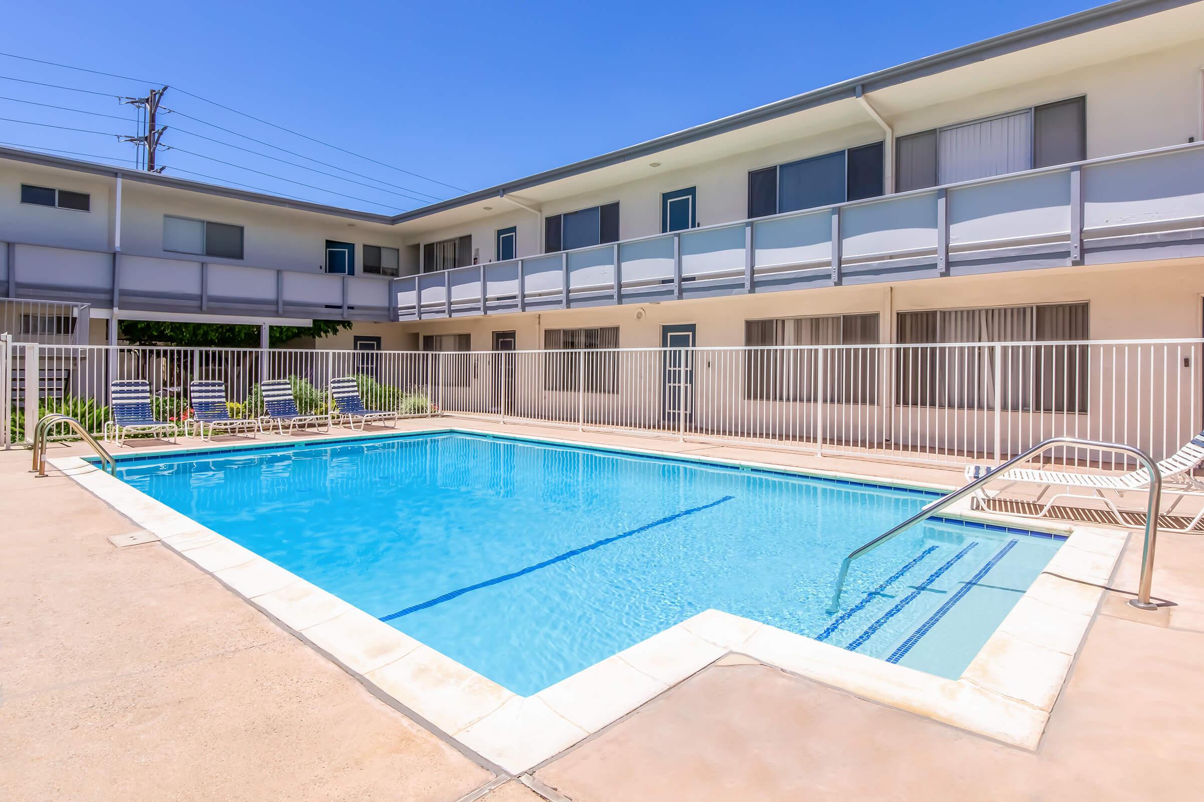 A clear blue swimming pool surrounded by a white fence, with lounge chairs positioned nearby. The pool area is framed by a light-colored building featuring multiple balconies. The sky is bright blue, indicating sunny weather, creating a relaxed and inviting atmosphere.