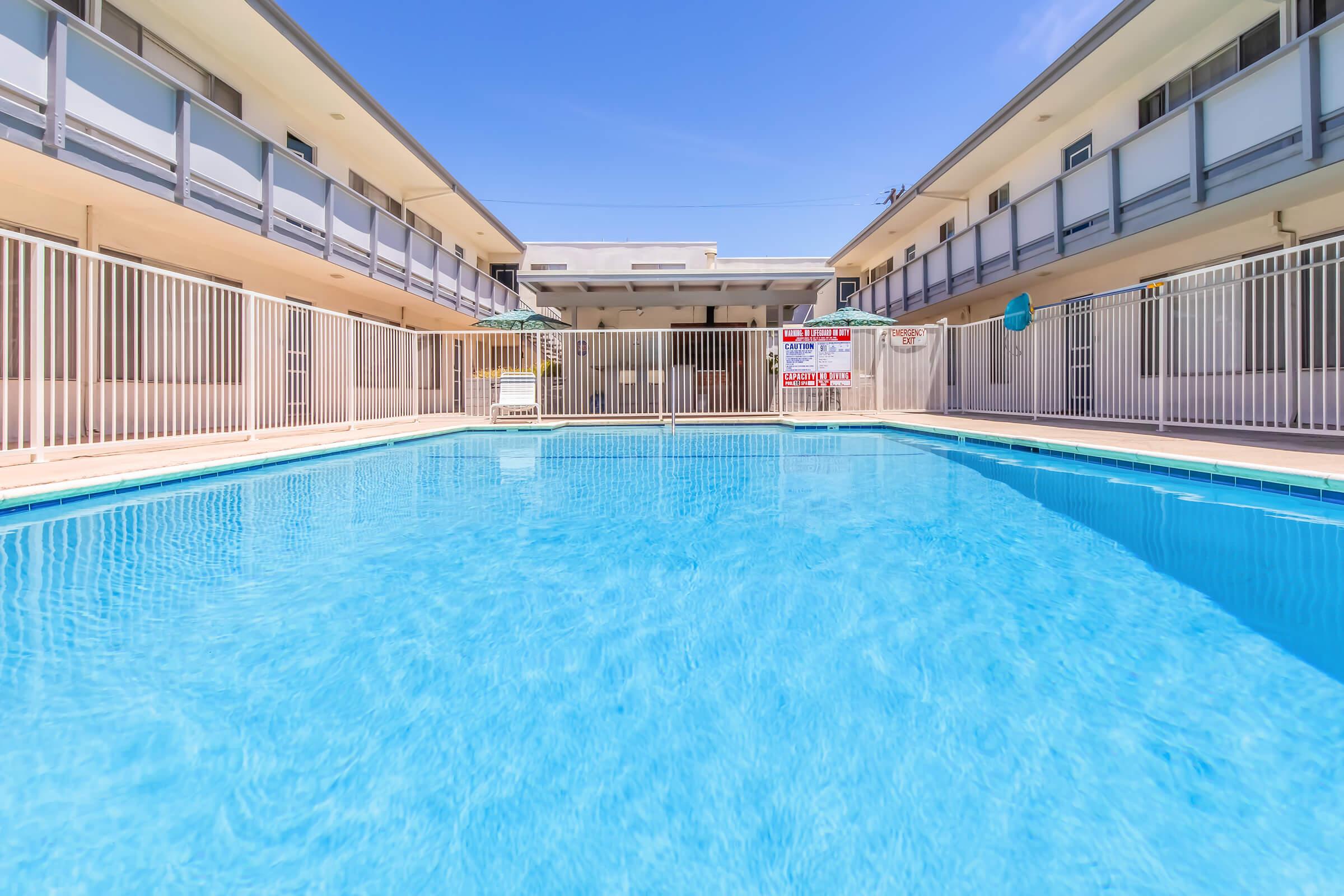 A clear blue pool surrounded by a white fence and two-story buildings. The sun shines brightly in a clear blue sky. Lounge chairs and umbrellas are positioned around the pool area, creating a relaxing atmosphere.