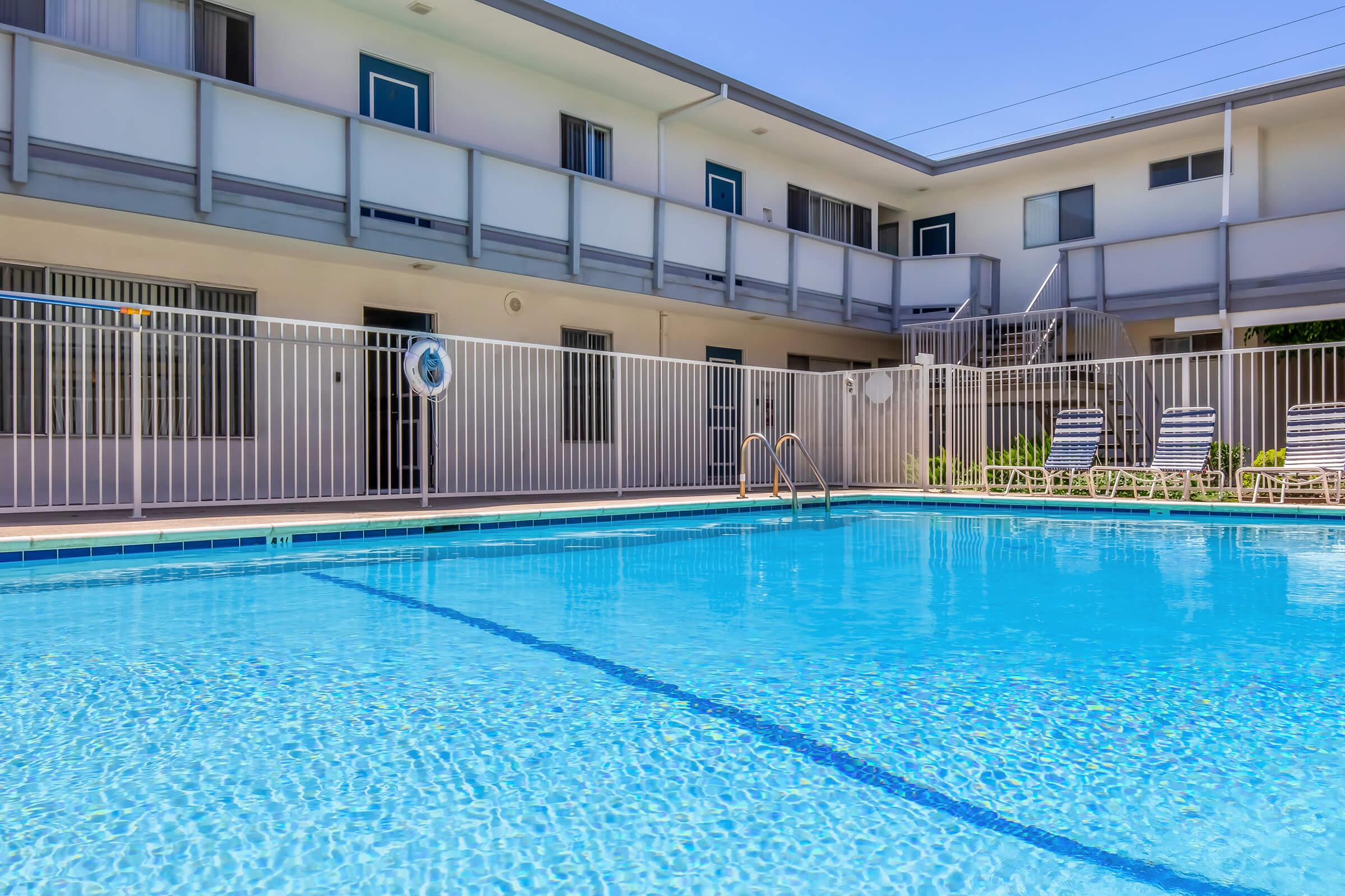A clear blue swimming pool surrounded by a white fence, with lounge chairs on the pool deck. In the background, a two-story building with multiple windows and doors can be seen, under a sunny sky. The scene conveys a relaxing atmosphere typical of a hotel or apartment complex.