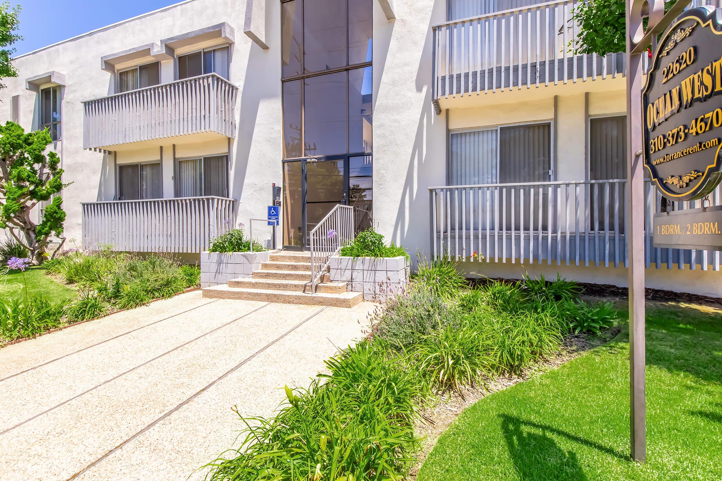 Exterior view of a modern apartment building with a glass entrance, featuring two levels of balconies. A pathway lined with low shrubs leads to the entrance. The building's address is displayed prominently. The landscaping includes green grass and blooming plants, creating a welcoming atmosphere.