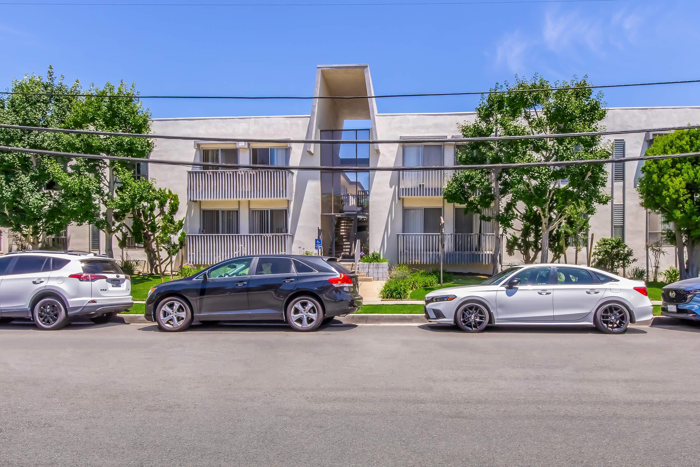 A modern apartment building with a unique architectural design, featuring large windows and a triangular entrance. Several cars are parked along the street in front, with trees providing greenery on either side. The sky is clear, indicating a sunny day.