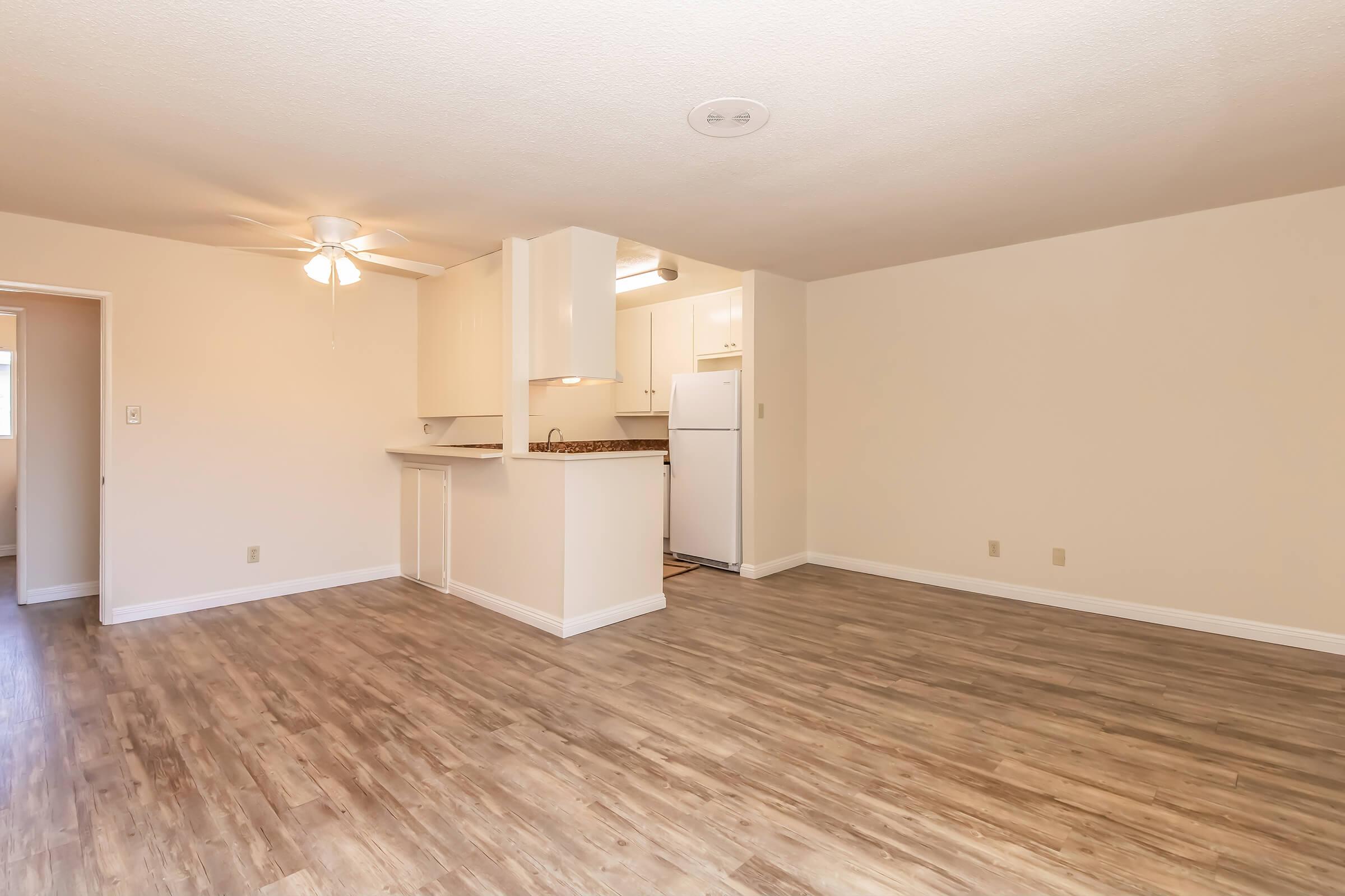 A spacious, well-lit living area featuring light-colored walls and wood-style flooring. To the left, there is an entryway leading to a kitchen with white cabinetry and appliances. A ceiling fan is visible, providing ventilation. The space is empty and ready for furniture arrangement.