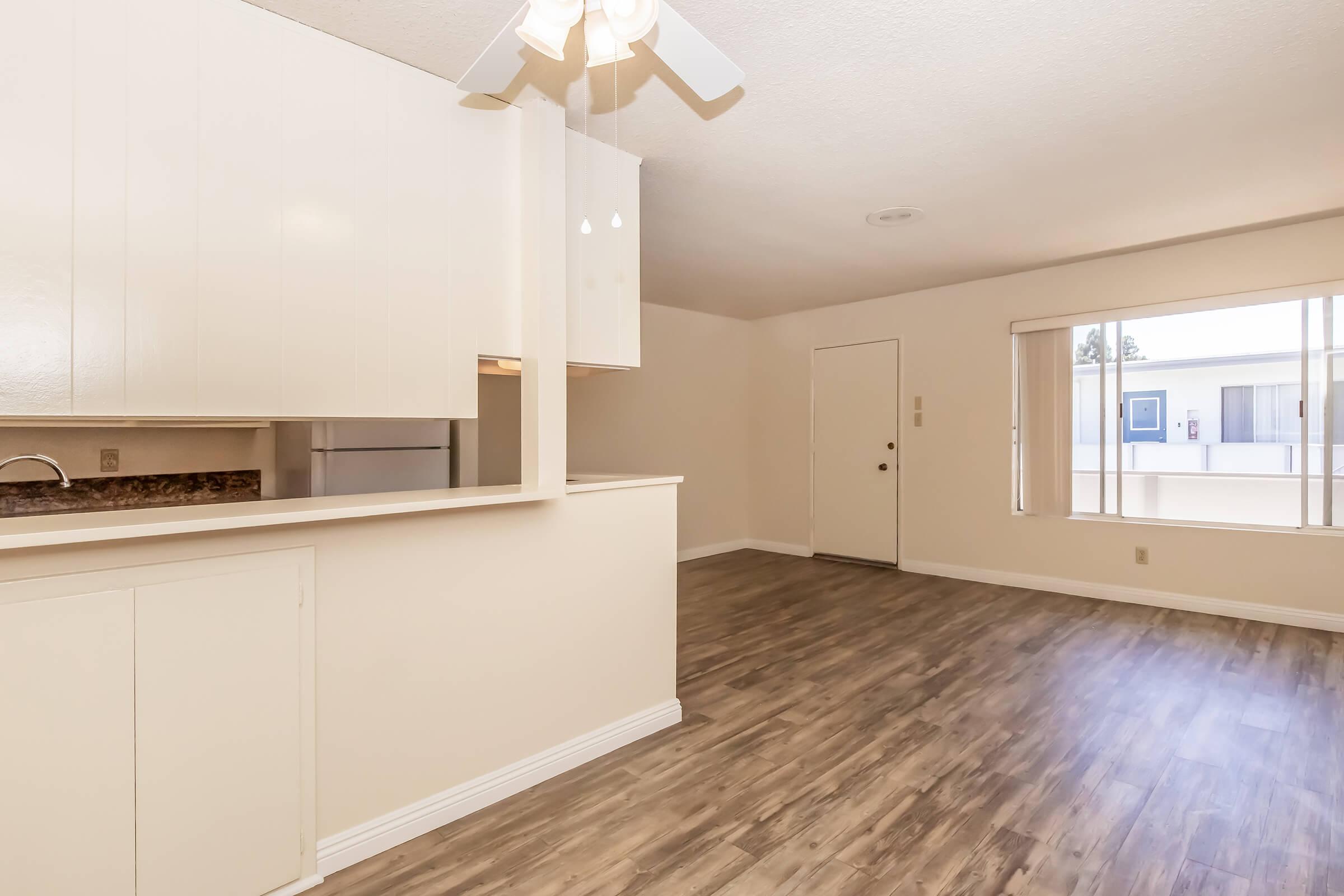 A spacious, well-lit living area featuring light-colored walls, modern wood-style flooring, and a ceiling fan. The open layout includes a kitchenette with a bar counter, and a large window allowing natural light. A door leads to an outside area.