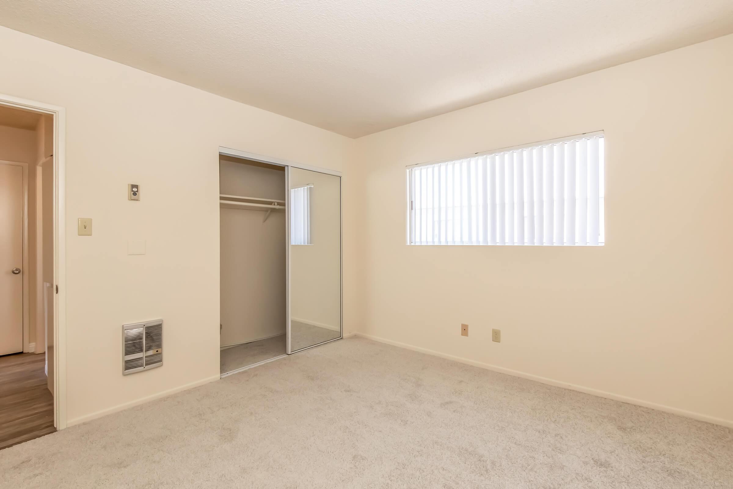 A vacant room with light-colored walls and a carpeted floor. It features a window with vertical blinds on one side, a closet with sliding doors, and a wall-mounted heater. The room is well-lit and has a clean, simple design, making it suitable for various uses.