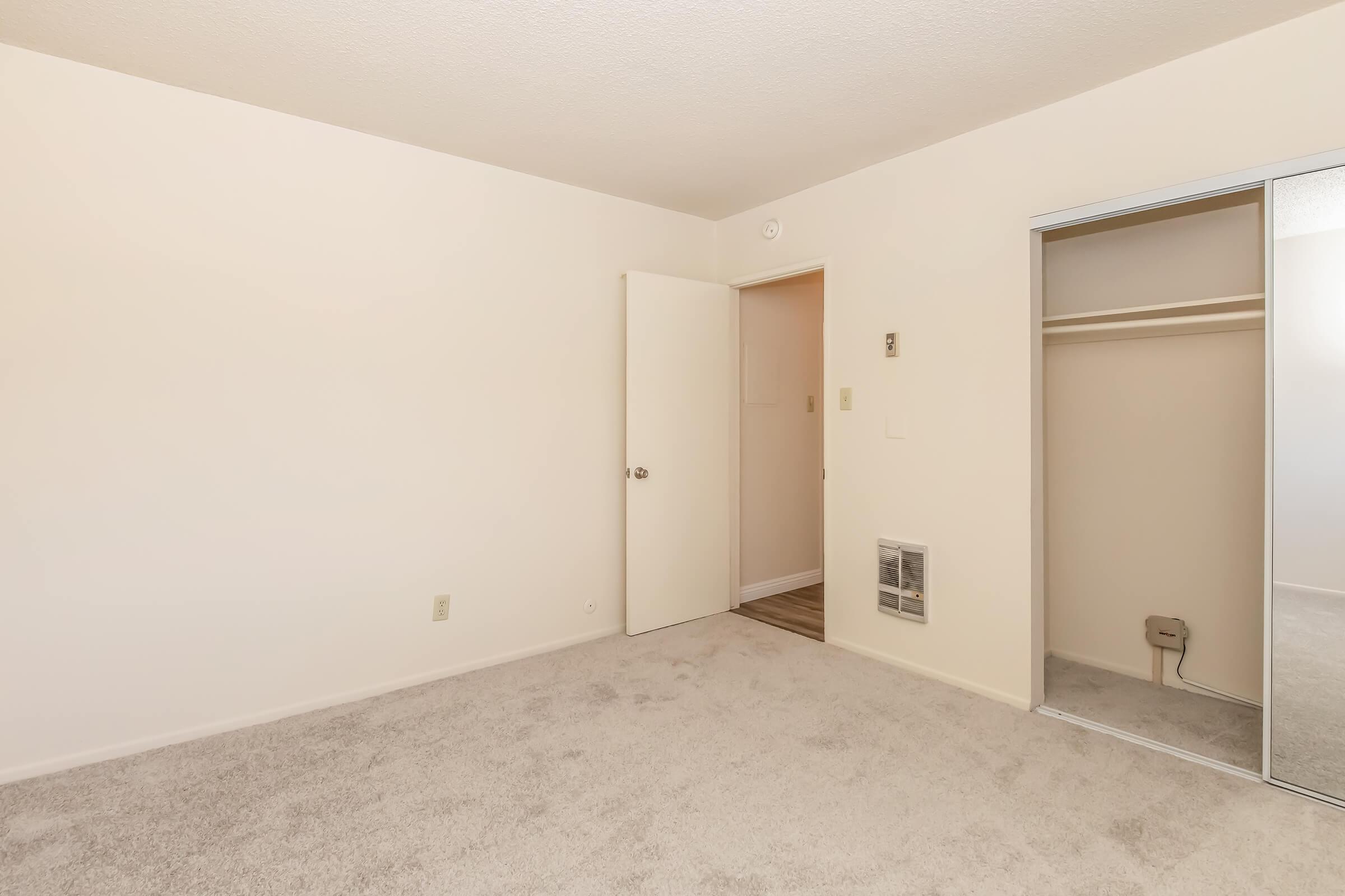 Empty room with light-colored walls and carpet. A closed door leads to another space, and there is a mirrored closet to the right. The floor is visible near the door, and a vent is located on the wall. The overall atmosphere is bright and uncluttered, suggesting a fresh, neutral living space.