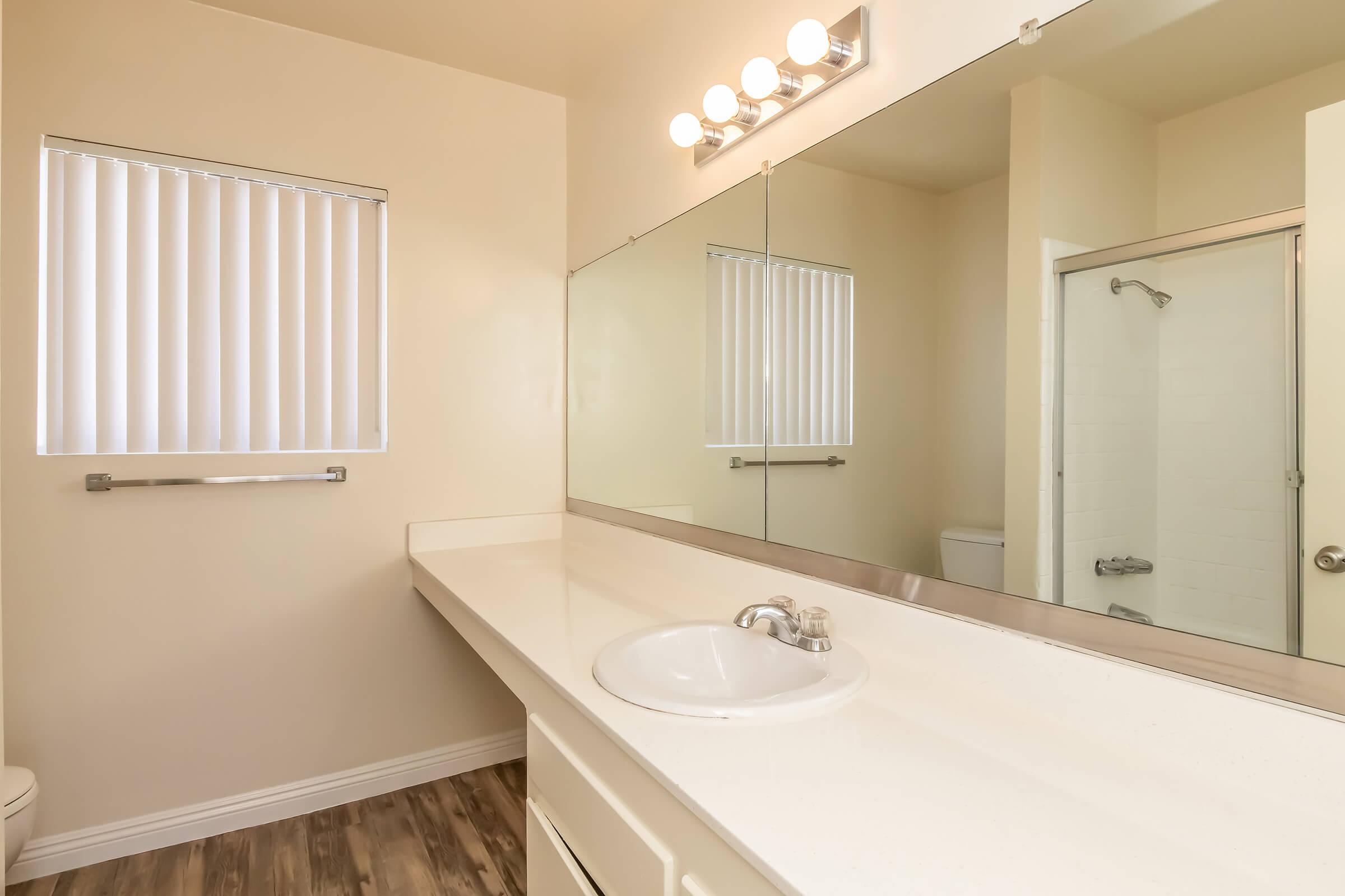 Bright and modern bathroom featuring a spacious countertop with a sink, a large mirror, and a shower area. Natural light enters through a window with vertical blinds. The flooring is wood-like, adding warmth to the space. The overall design is clean and minimalistic.