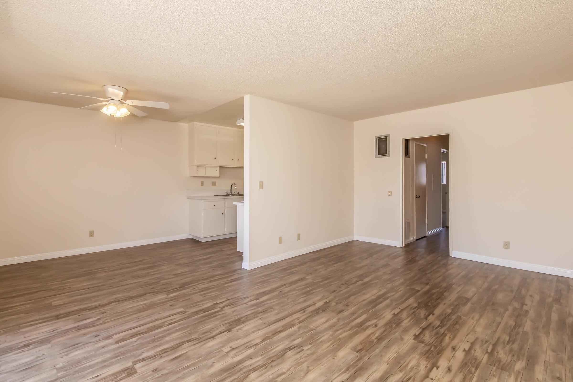A spacious, well-lit living area featuring light-colored walls and a ceiling fan. The floor is finished with laminate wood, and a doorway leads to adjacent rooms. A small kitchenette is visible in the background on the left, equipped with white cabinets. The overall ambiance is open and inviting.