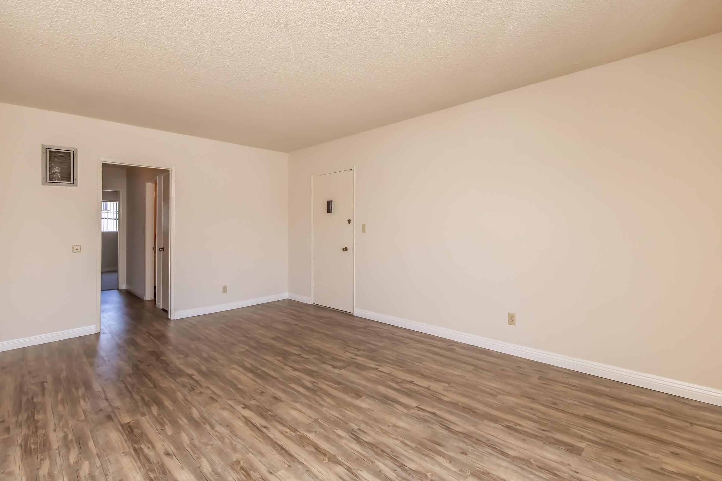 A spacious, empty room with light-colored walls and a wood-like floor. There's a doorway on the left leading to another room and a door on the right. The room has no furniture, creating a bright and airy atmosphere.