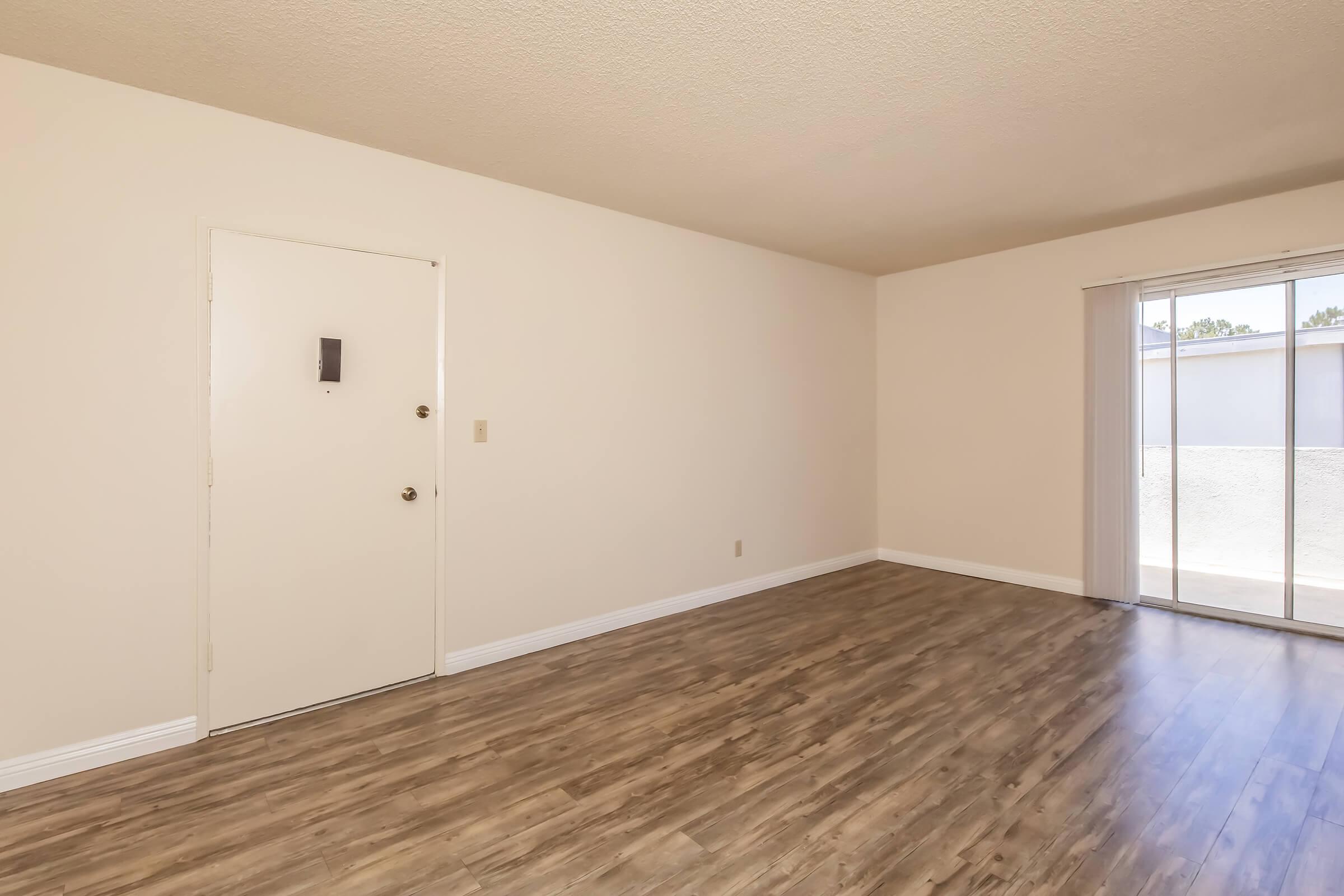 A spacious, empty room with light-colored walls and a large sliding glass door leading to a balcony. The floor is covered with light brown laminate, creating a warm atmosphere. A simple white door is visible on one wall, and there is a small wall switch nearby. The room is well-lit with natural light.