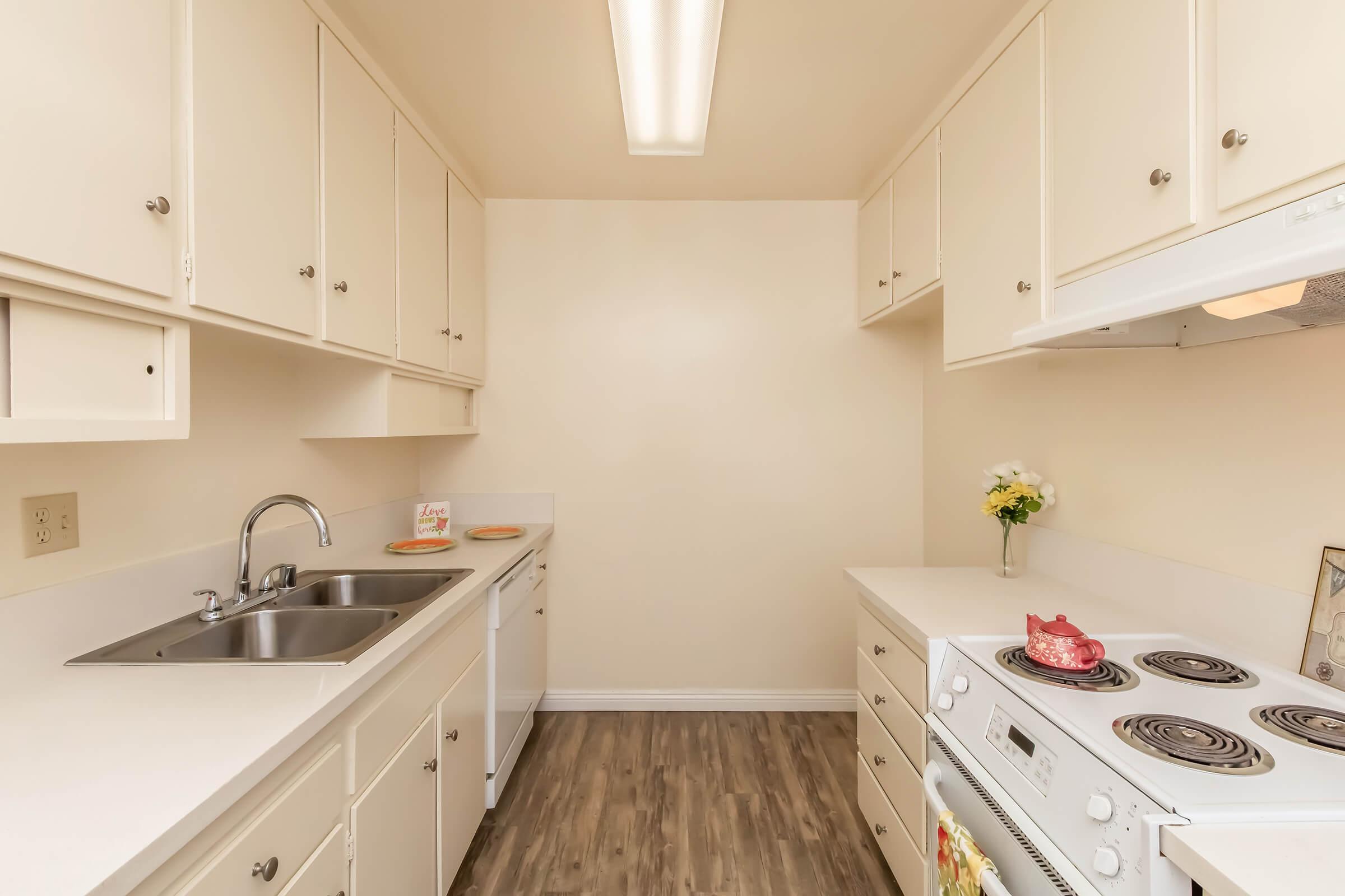 A small, modern kitchen featuring white cabinetry, a double sink, a dishwasher, and a stove with a vent. The countertops are light-colored, and there are decorative elements like a vase with flowers and plates on the counter. The flooring is a wood-like laminate, creating a cozy atmosphere.