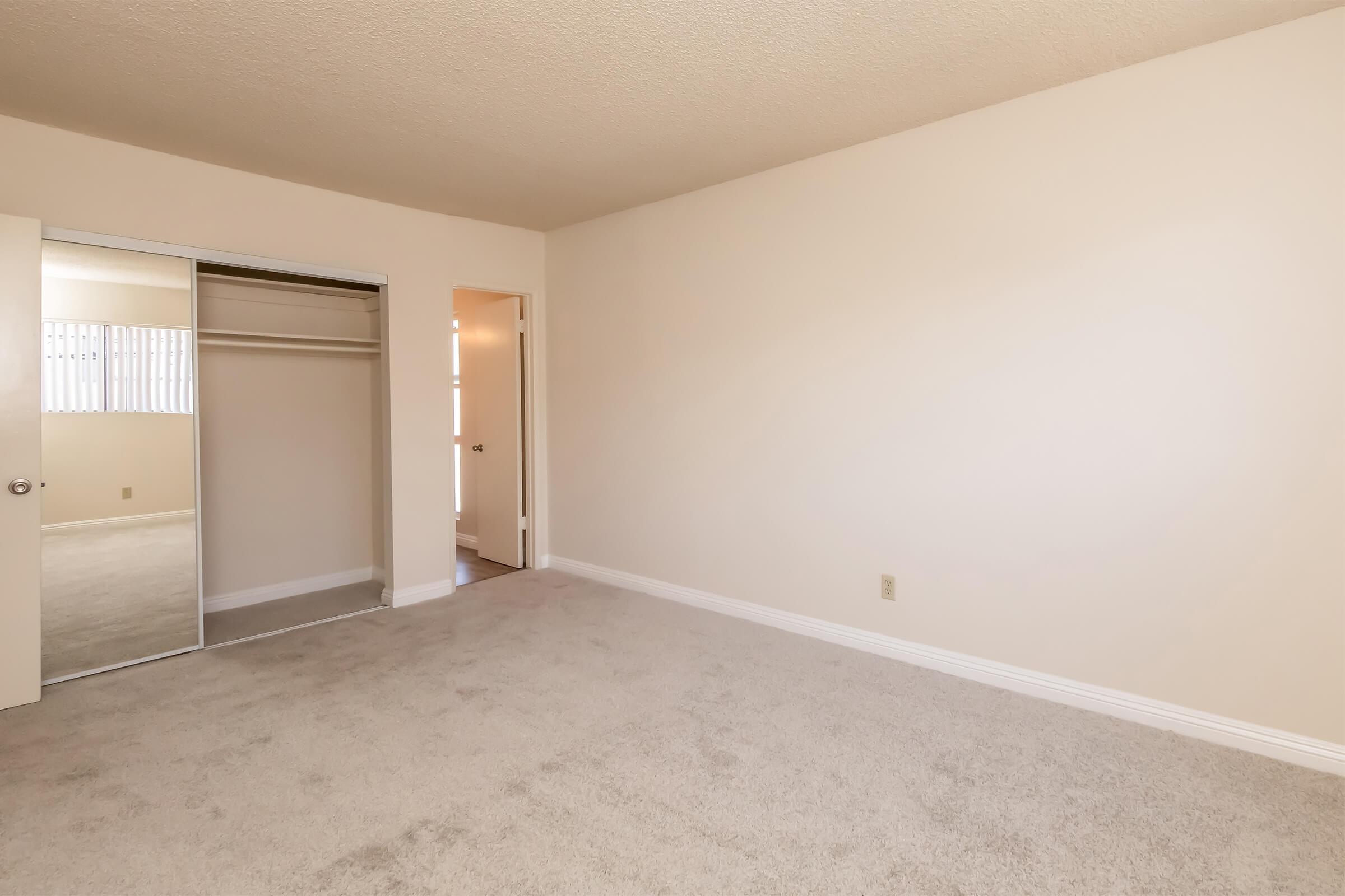 A spacious, empty bedroom with beige walls and light carpet. There's a mirrored closet door on the left, and a doorway leading to another room on the right. The room is well-lit with natural light coming from a window in the distance.