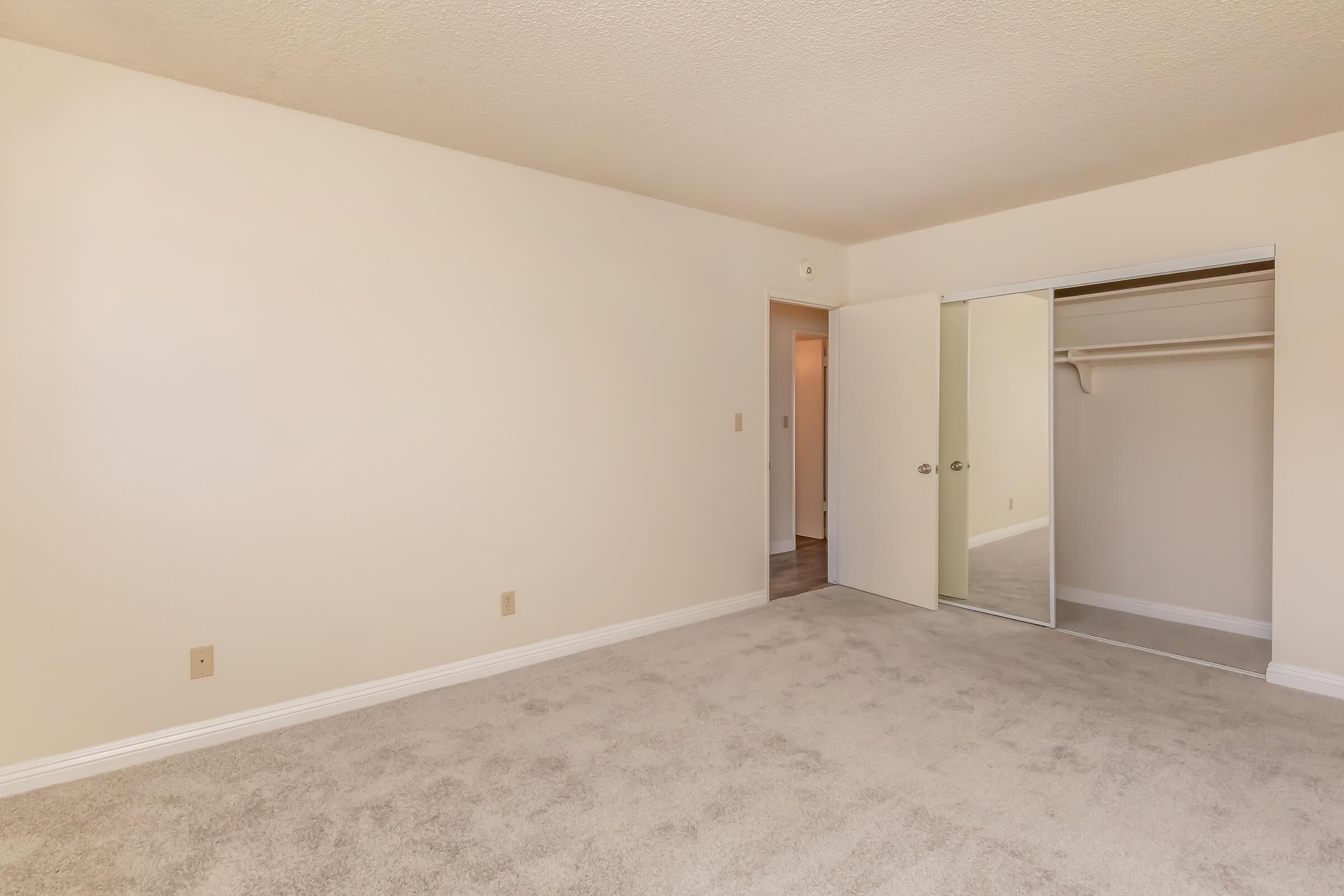 A spacious, empty room with light beige walls and a plush, carpeted floor. There is a mirrored closet door on the left, leading to a simple closet space. The room appears well-lit and inviting, with a doorway visible in the background, suggesting access to another space.