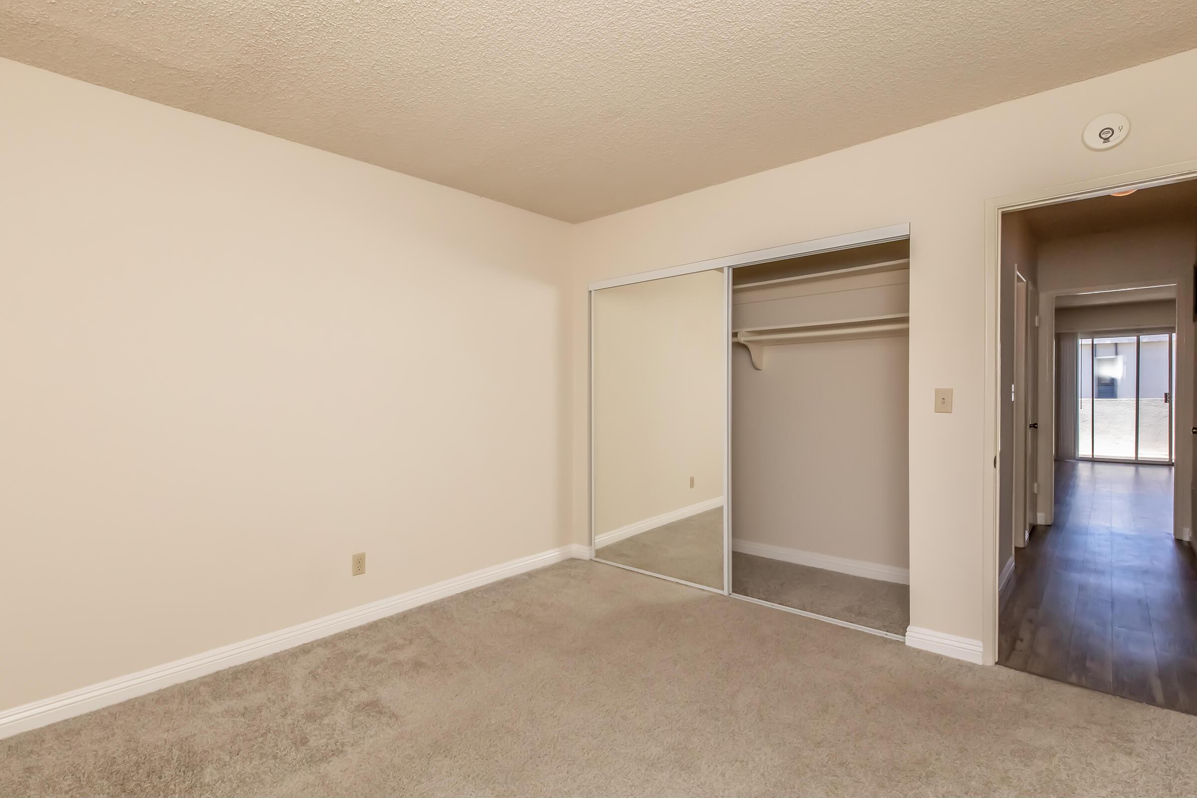 Empty room with beige walls, carpeted floor, and mirrored closet doors. A hallway is visible in the background leading to a sliding glass door. Natural light filters into the room, creating a bright and open atmosphere.