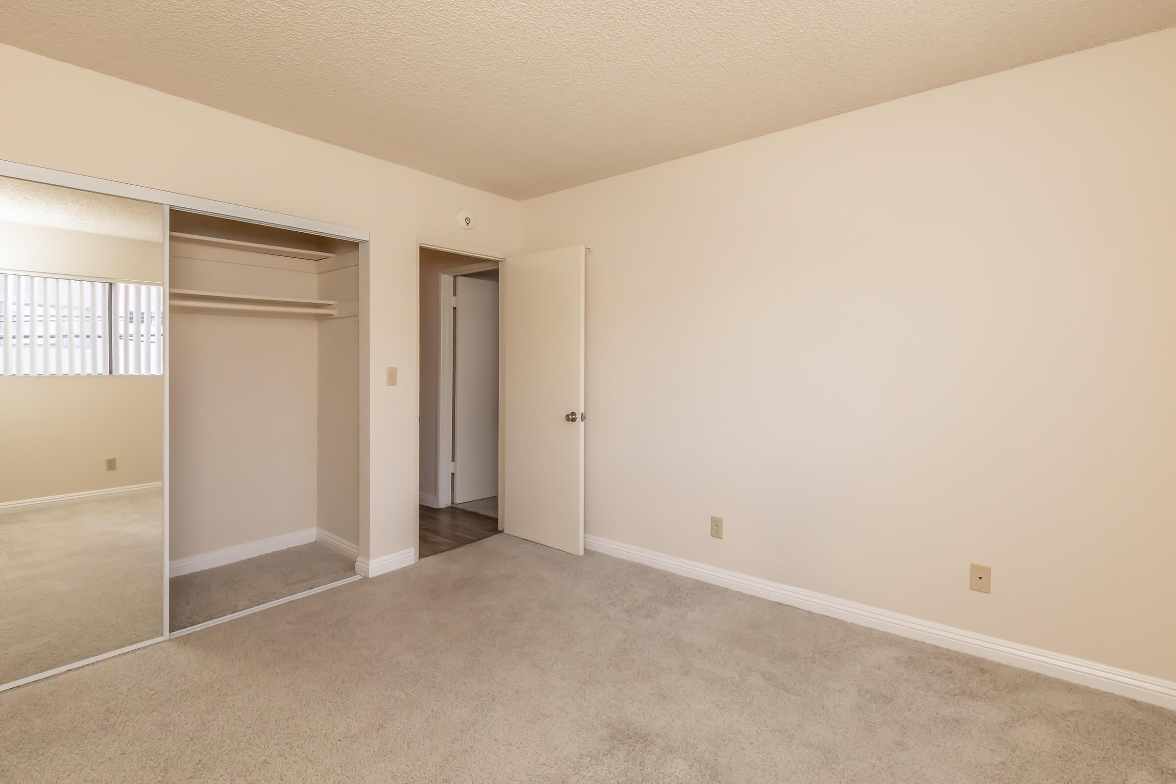 Empty room with light beige walls and carpet. A mirrored closet is on the left, showing its interior. There is a door leading to another space visible in the background, with natural light coming from the windows behind the closet. The overall aesthetic is clean and simple.