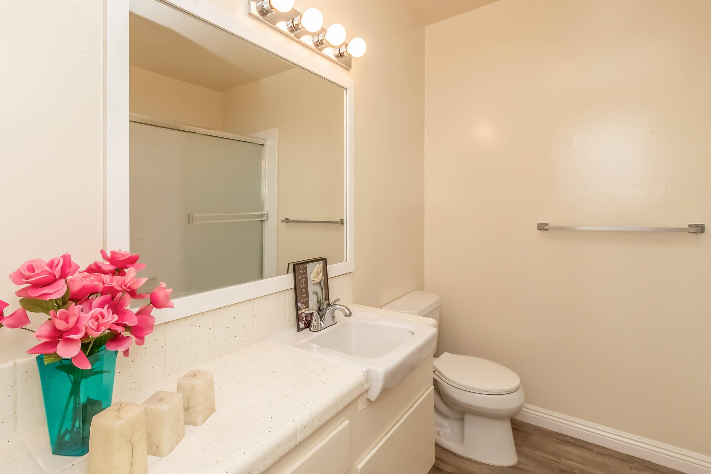 A bright, modern bathroom featuring a large mirror with five light fixtures above. There is a white sink and toilet, a shower behind a glass door, and a countertop adorned with pink flowers in a turquoise vase. Two white candles sit on the countertop, adding a decorative touch. The walls are painted a light color.