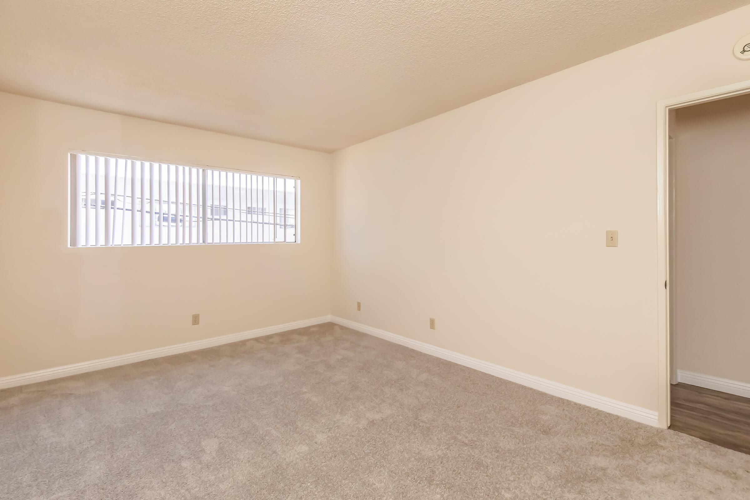 A spacious, empty room with beige walls and light gray carpet. A window with vertical blinds allows natural light to enter, and a door on the right leads to another area. The room has a clean, simple design, suitable for various uses such as a living room or office.