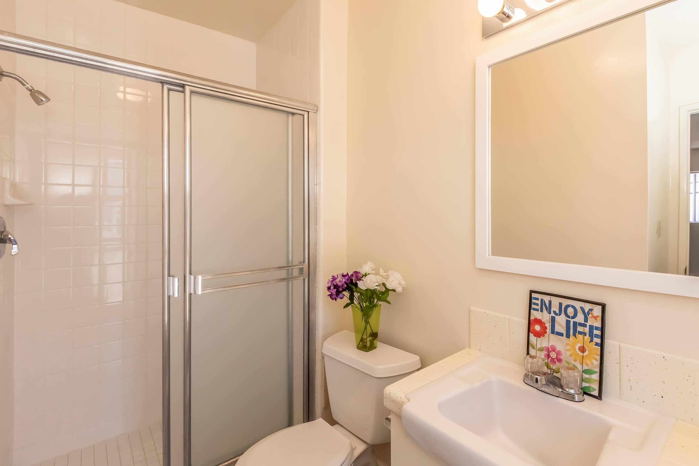 A clean, modern bathroom featuring a walk-in shower with clear glass doors, a white toilet, and a sink with a countertop. A vase with flowers adds a decorative touch, and a framed sign that reads "ENJOY LIFE" is displayed on the counter. The walls are light-colored, contributing to a bright and airy atmosphere.