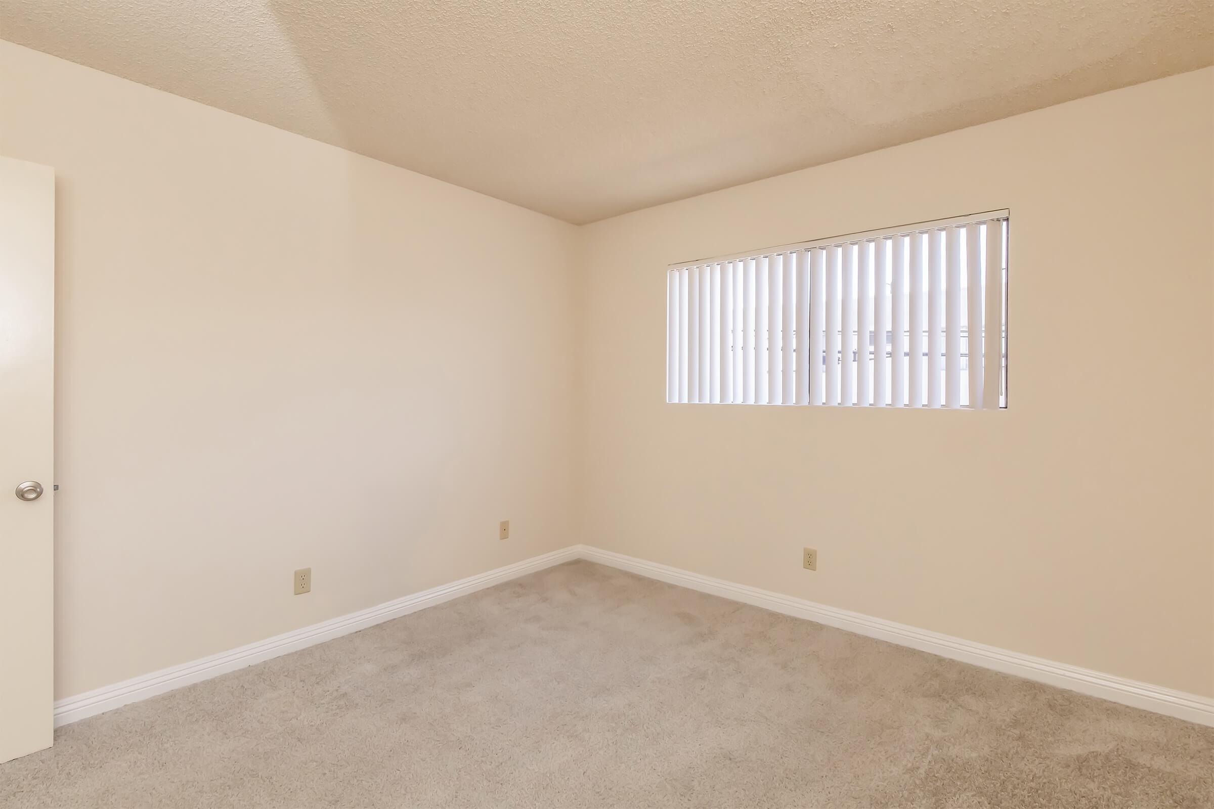 Empty room with beige walls and carpeted floor, featuring a single window with vertical blinds. There are light fixtures on the ceiling and minimal wall detailing.