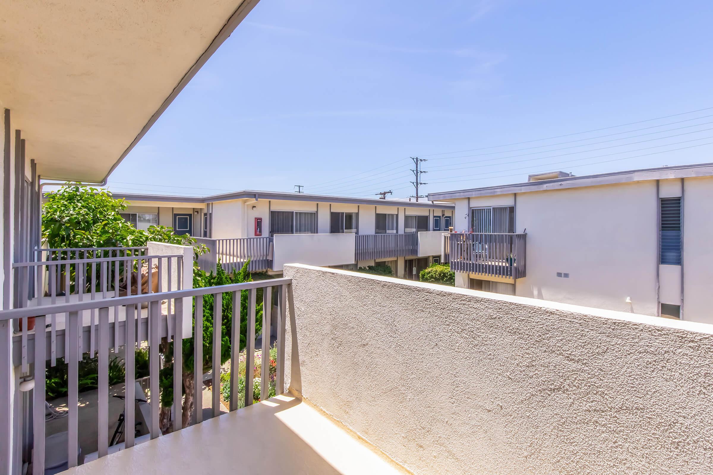 View from a balcony overlooking a courtyard surrounded by apartment buildings. The scene includes plants and trees, with clear blue skies in the background. The buildings are two stories high, displaying balconies and windows, creating a residential atmosphere.