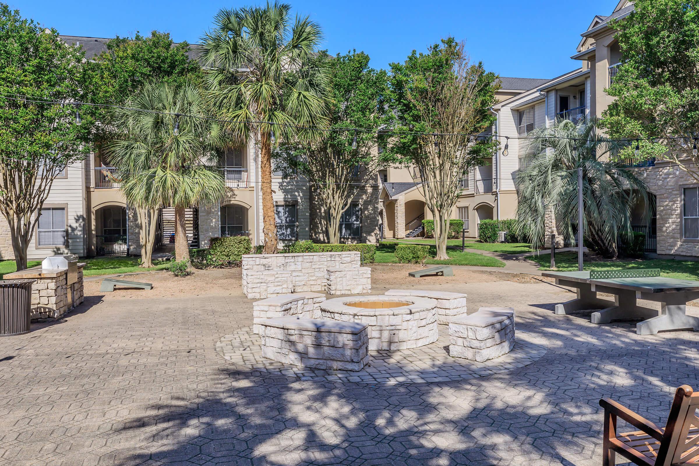 A landscaped courtyard featuring stone fire pits surrounded by seating areas, lush palm trees, and well-maintained greenery. The setting includes several residential buildings in the background, with clear blue skies overhead, creating a welcoming outdoor space.
