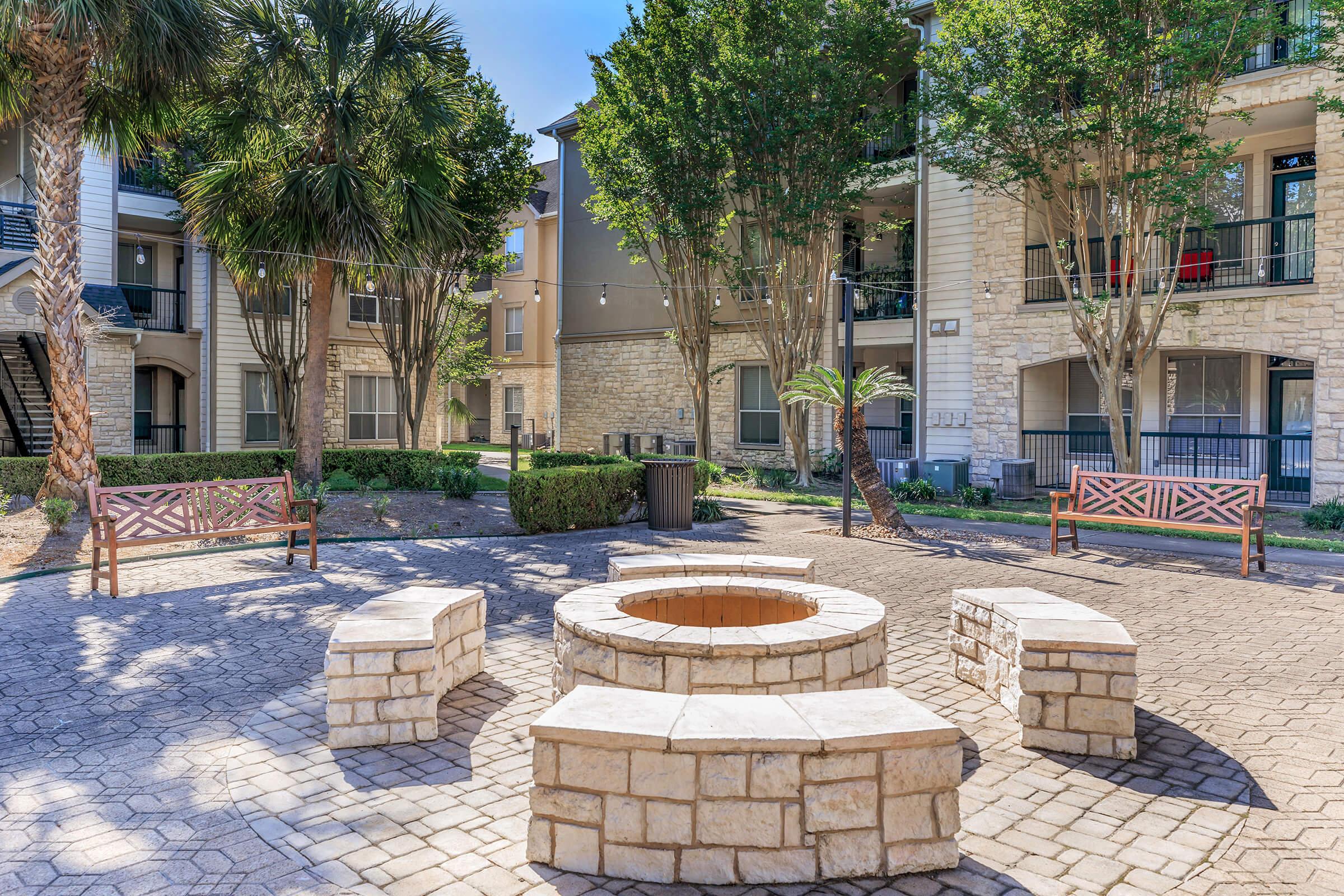 A circular stone fire pit surrounded by five stone benches, situated in a landscaped courtyard with palm trees and nearby apartment buildings. The area is sunny and features greenery, creating a welcoming outdoor space for gatherings.