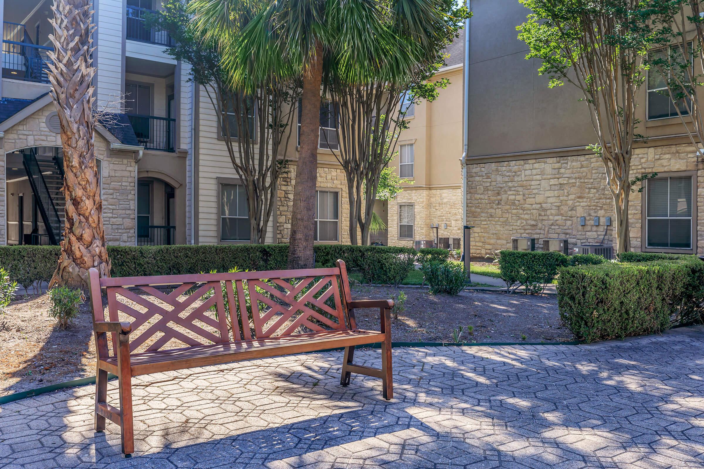 A wooden bench with a geometric design sits on a paved path in a landscaped area surrounded by trees and manicured hedges. The background features apartment buildings with a mix of brick and stucco exteriors, creating a calm and inviting outdoor space.