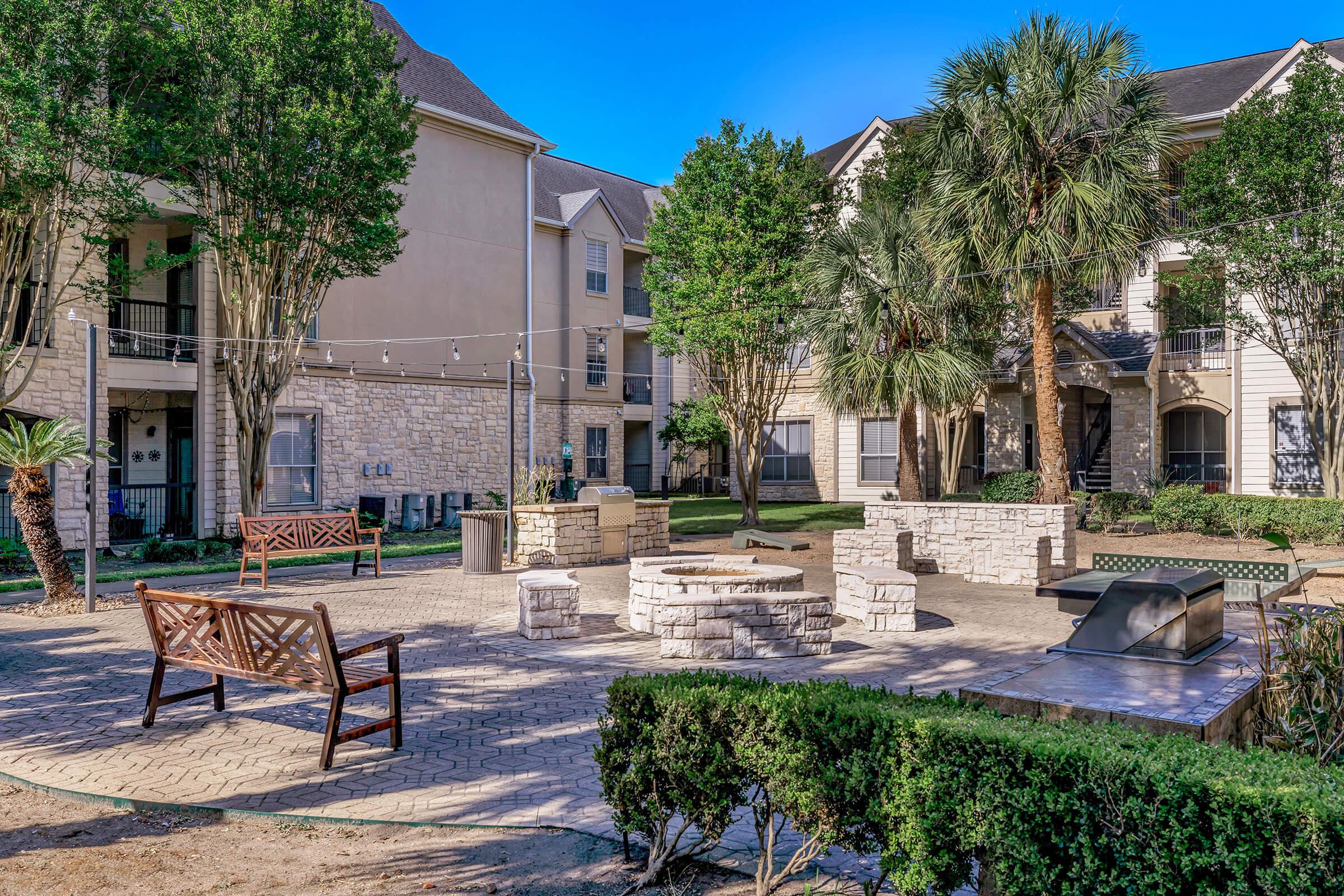 A landscaped outdoor common area featuring stone fire pits, wooden benches, and palm trees. The surroundings include modern apartment buildings with greenery, creating a peaceful, communal atmosphere for residents. Bright blue sky overhead enhances the inviting scene.
