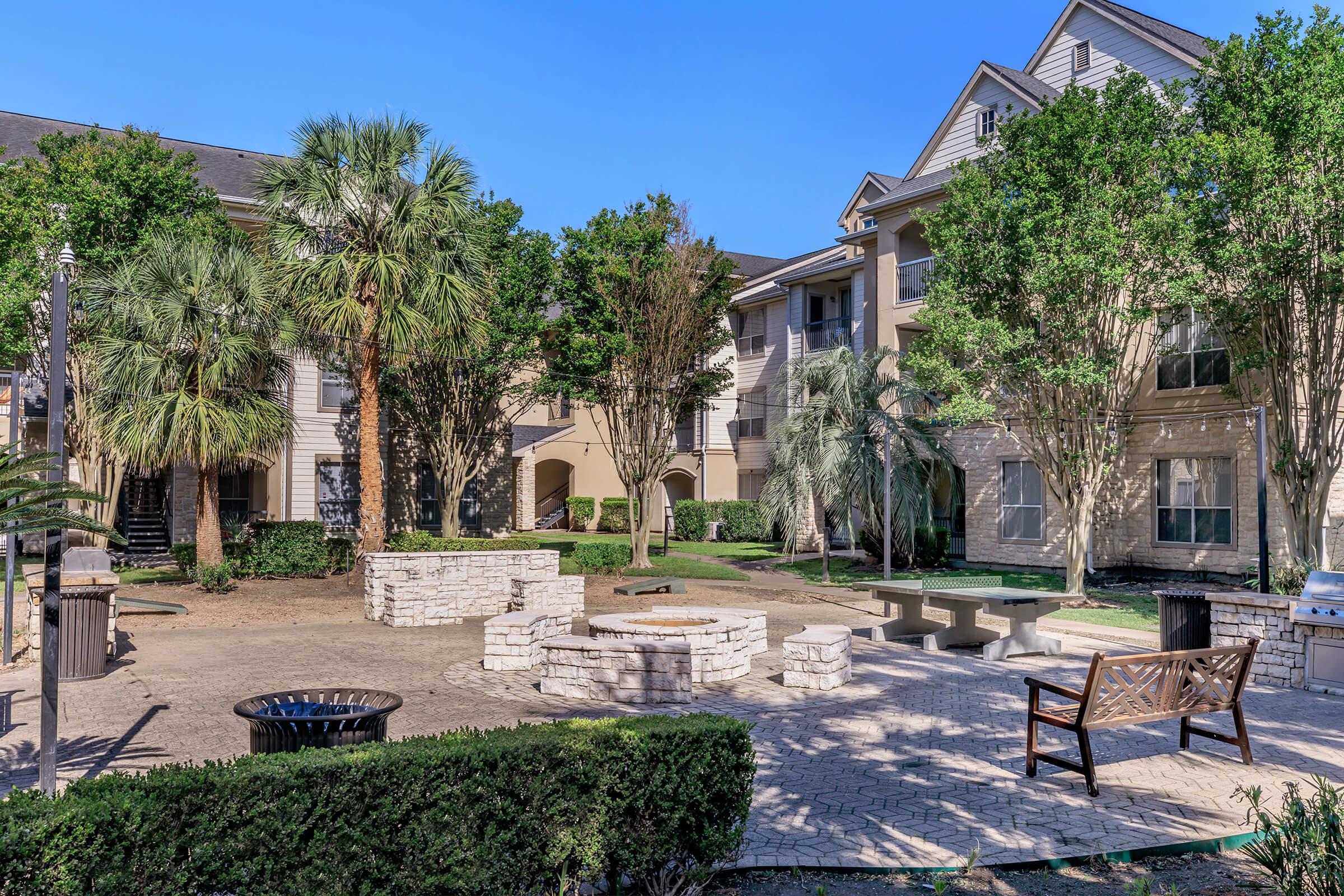 A well-maintained courtyard featuring palm trees, green shrubs, and a stone fire pit. The area includes seating options such as benches and a BBQ grill, surrounded by modern apartment buildings under a clear blue sky. Ideal for relaxation and outdoor gatherings.