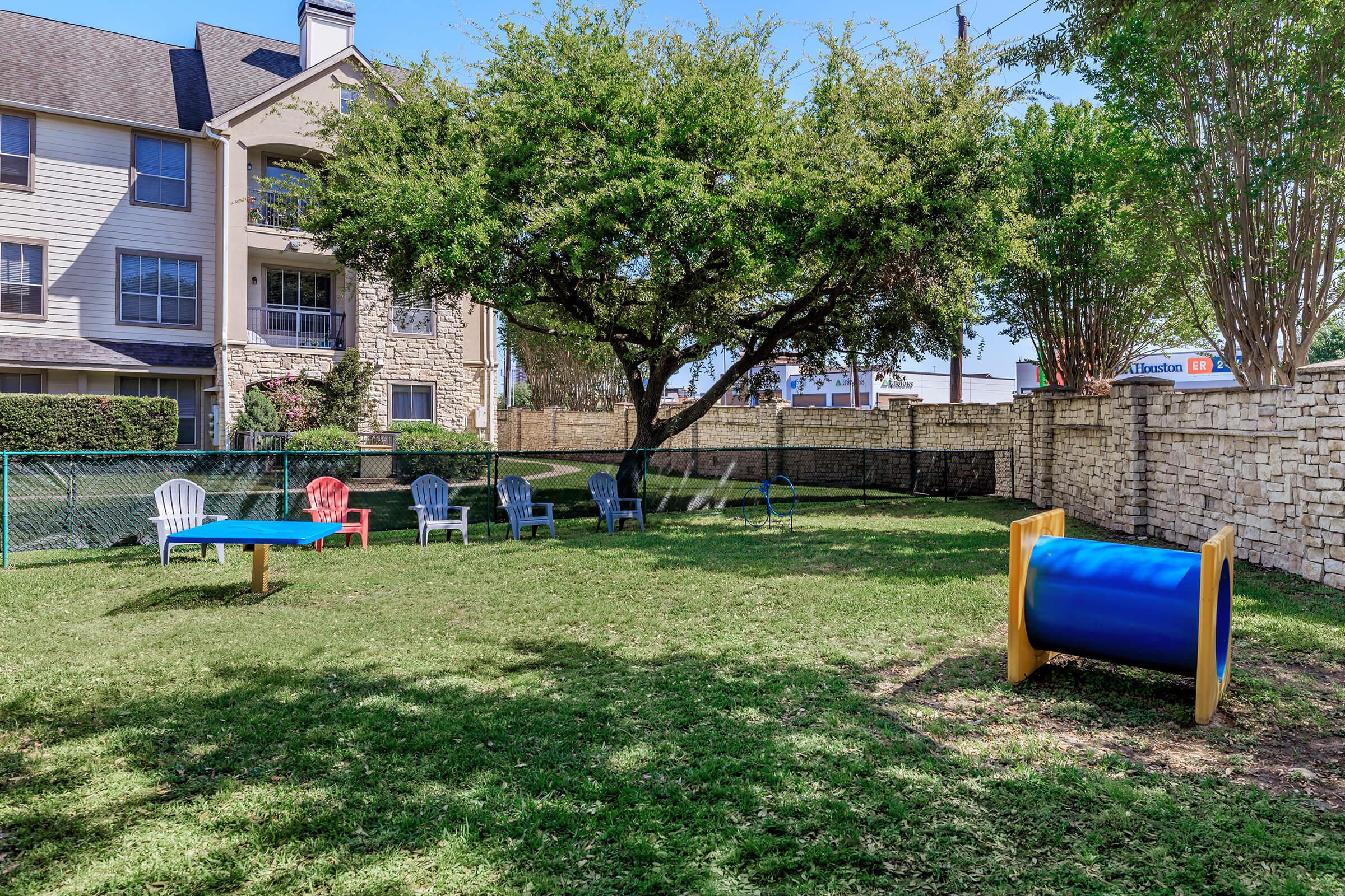 A grassy outdoor area featuring several colorful chairs, a blue barrel-shaped tunnel, and a wooden table. The background includes an apartment building and a fenced perimeter with trees providing shade.