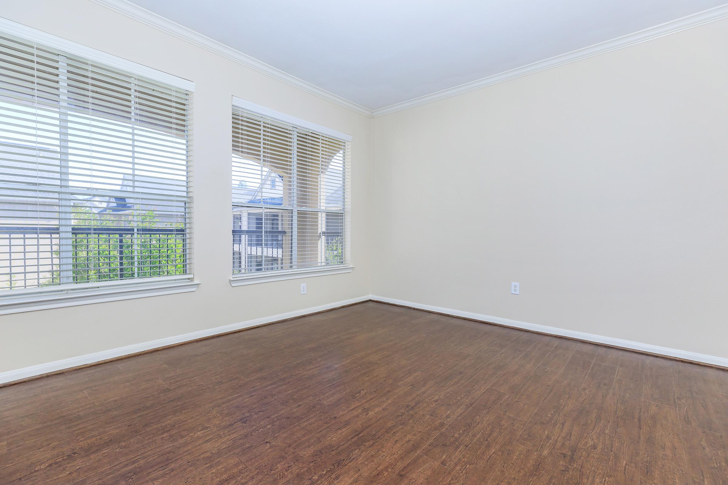 A vacant room with light-colored walls and wooden flooring. There are two large windows with blinds allowing natural light to enter, and no furniture is present in the space. The overall atmosphere is bright and open, creating a blank canvas suitable for various uses.